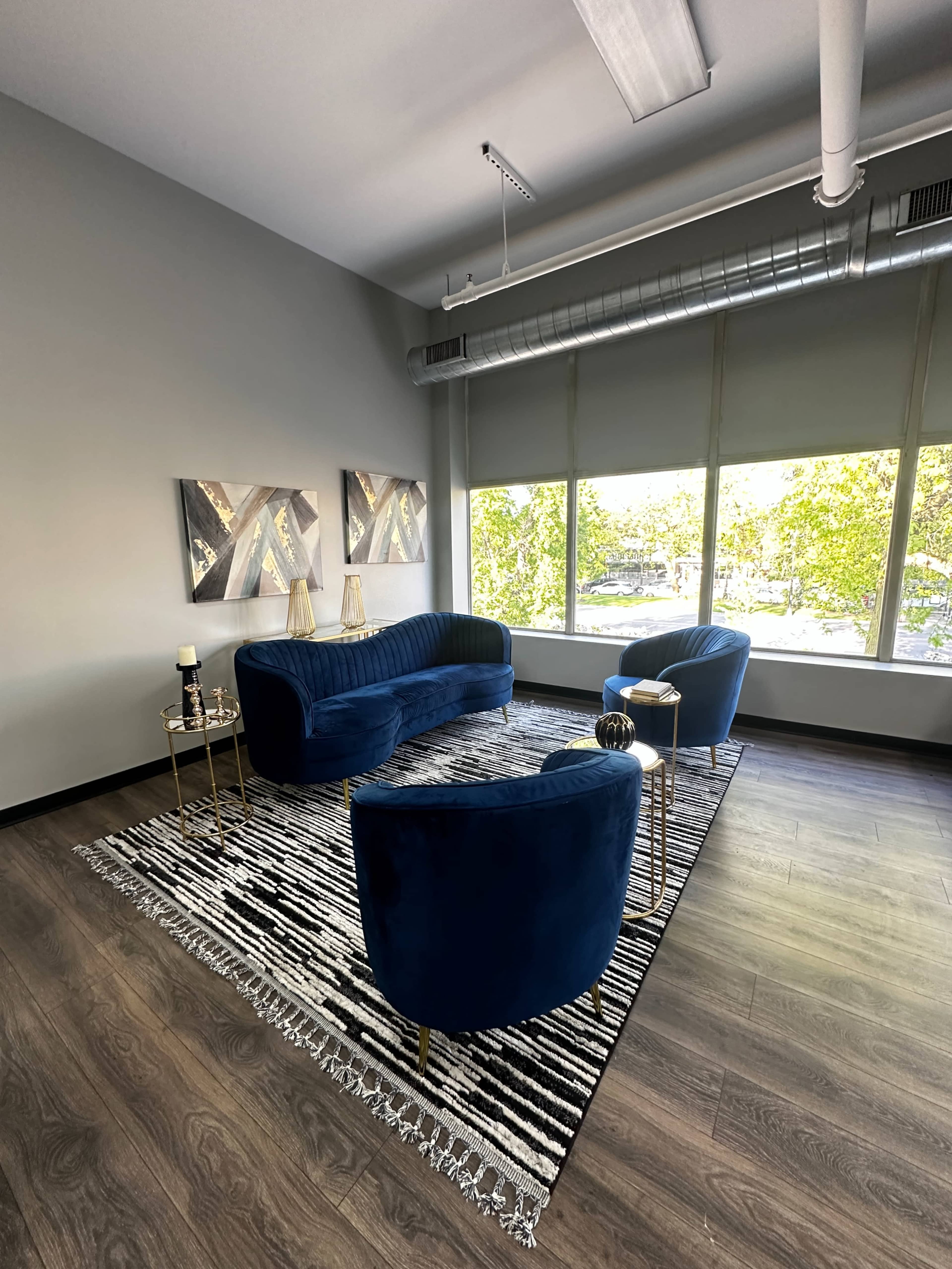 The image shows a modern seating area with a blue velvet sofa and two matching chairs arranged around a striped rug under natural light from large windows.