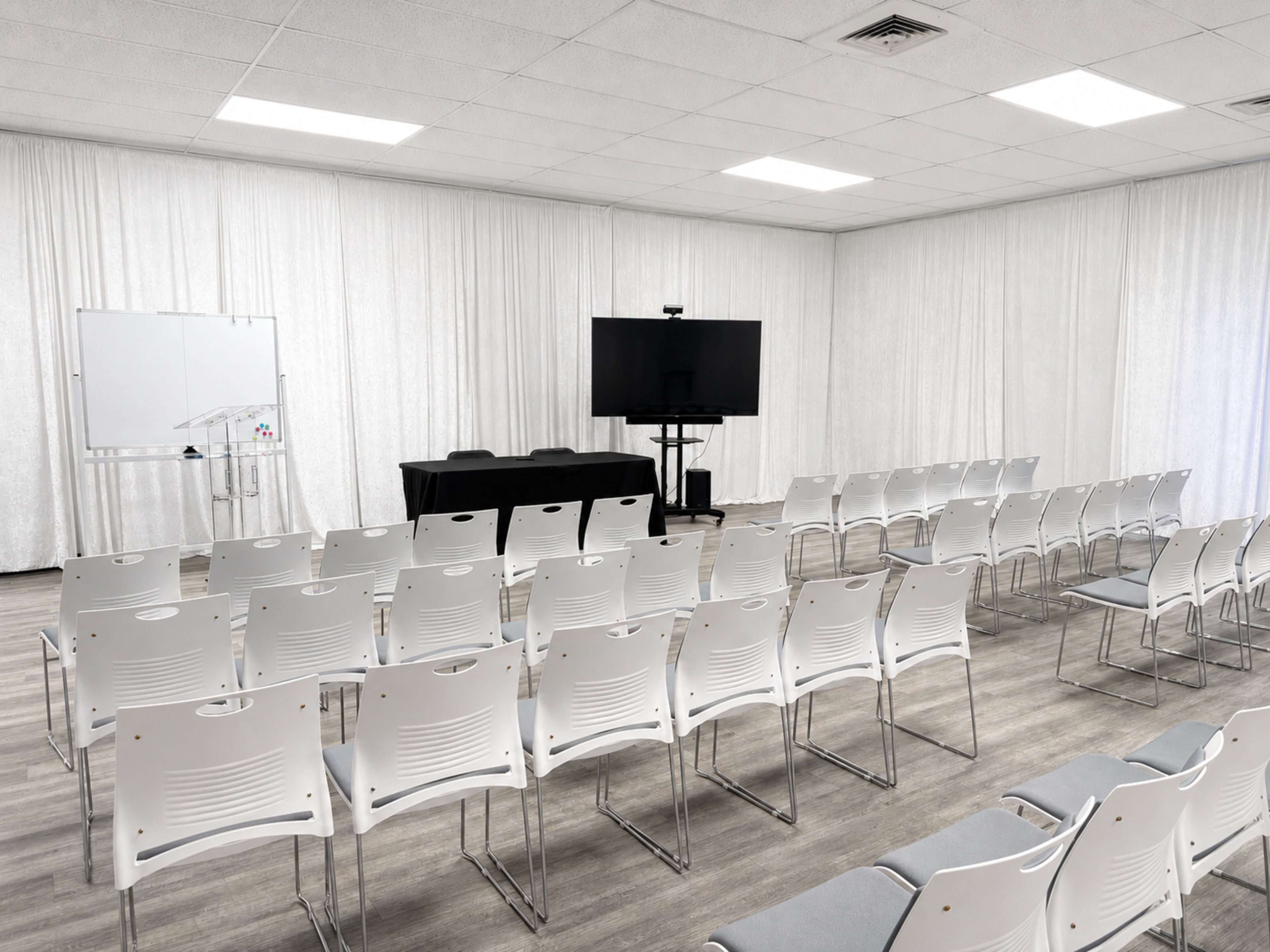 A conference room is set up with rows of white chairs facing a black table and a large screen, with a whiteboard positioned nearby.
