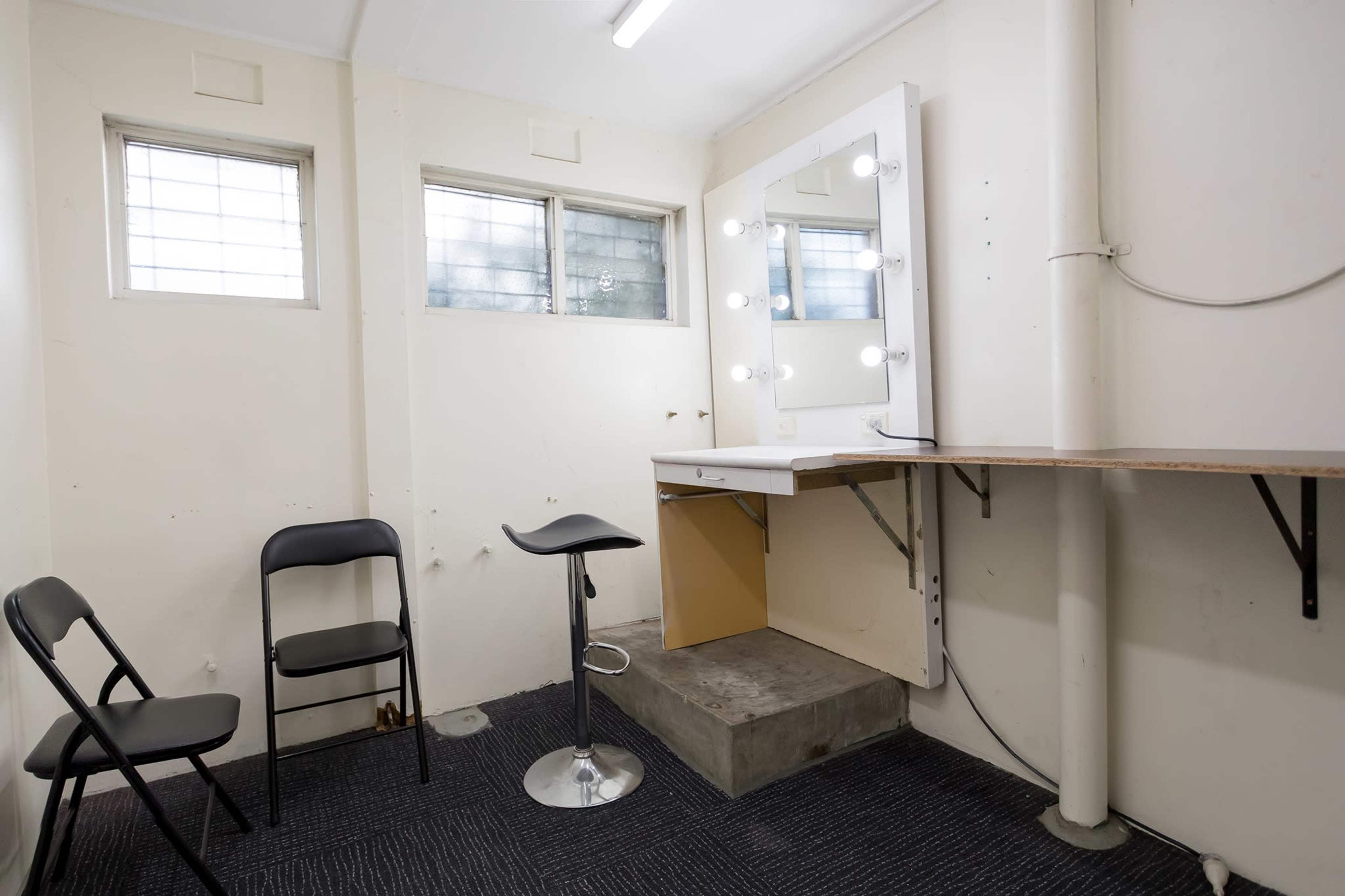 A sparse room with a makeup station featuring illuminated mirrors, a stool, and two collapsible chairs against the wall.