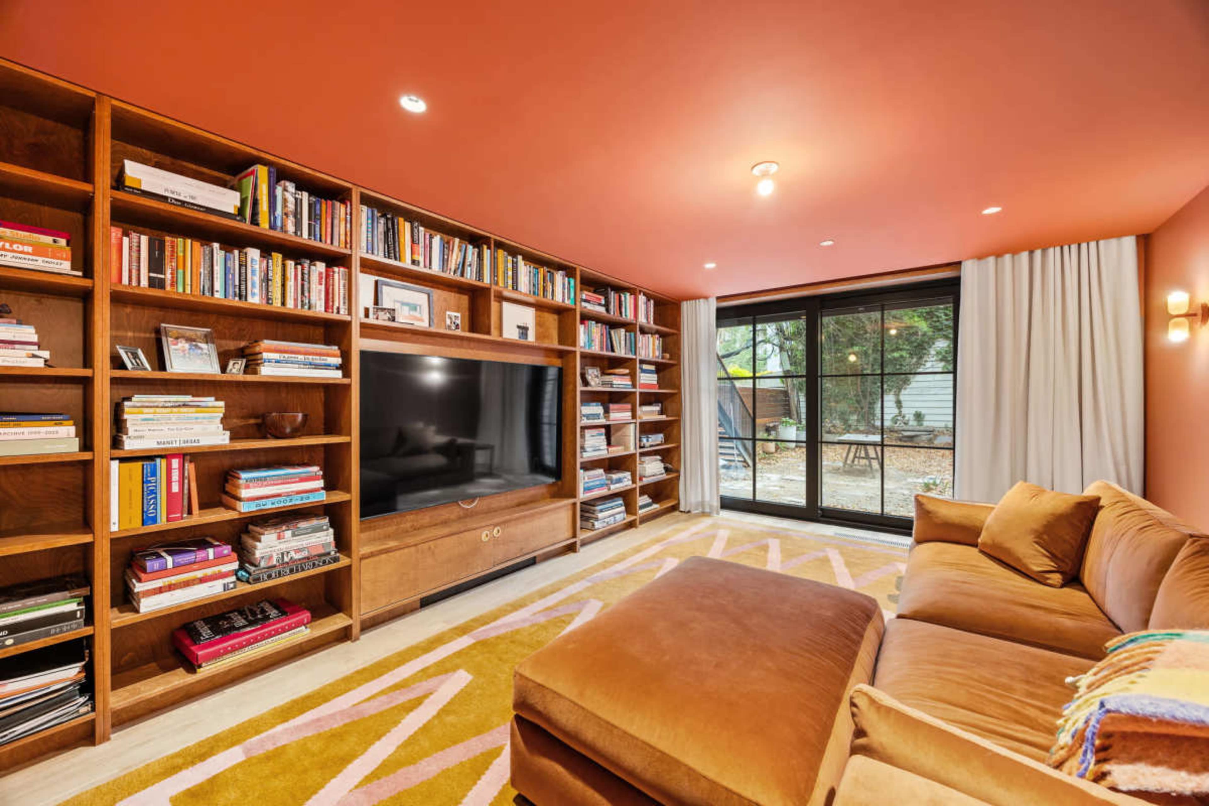 The image shows a cozy living room featuring a large bookcase filled with books, a television, and a brown couch with a colorful rug on the floor, alongside a large window with curtains.
