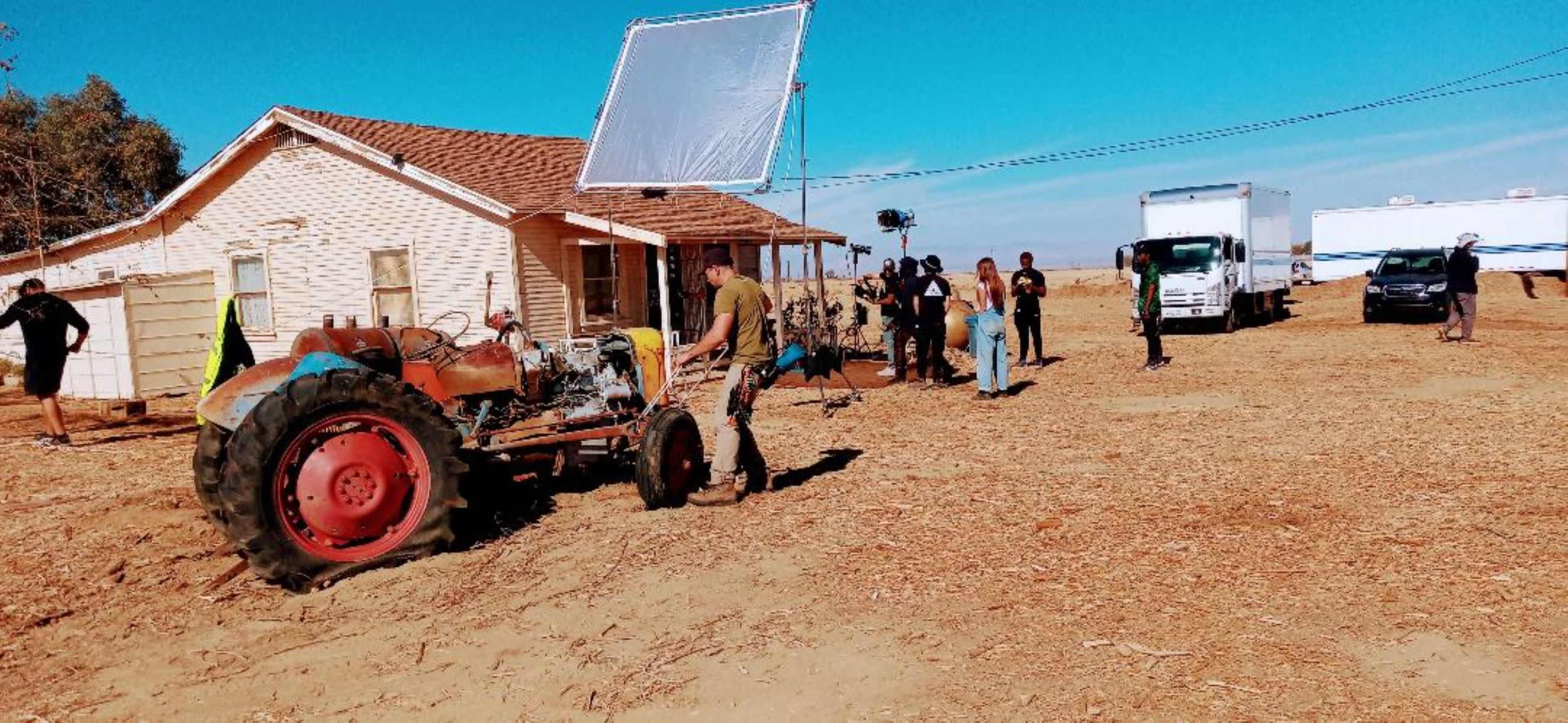 A film crew sets up equipment around a tractor and a house in a rural area.