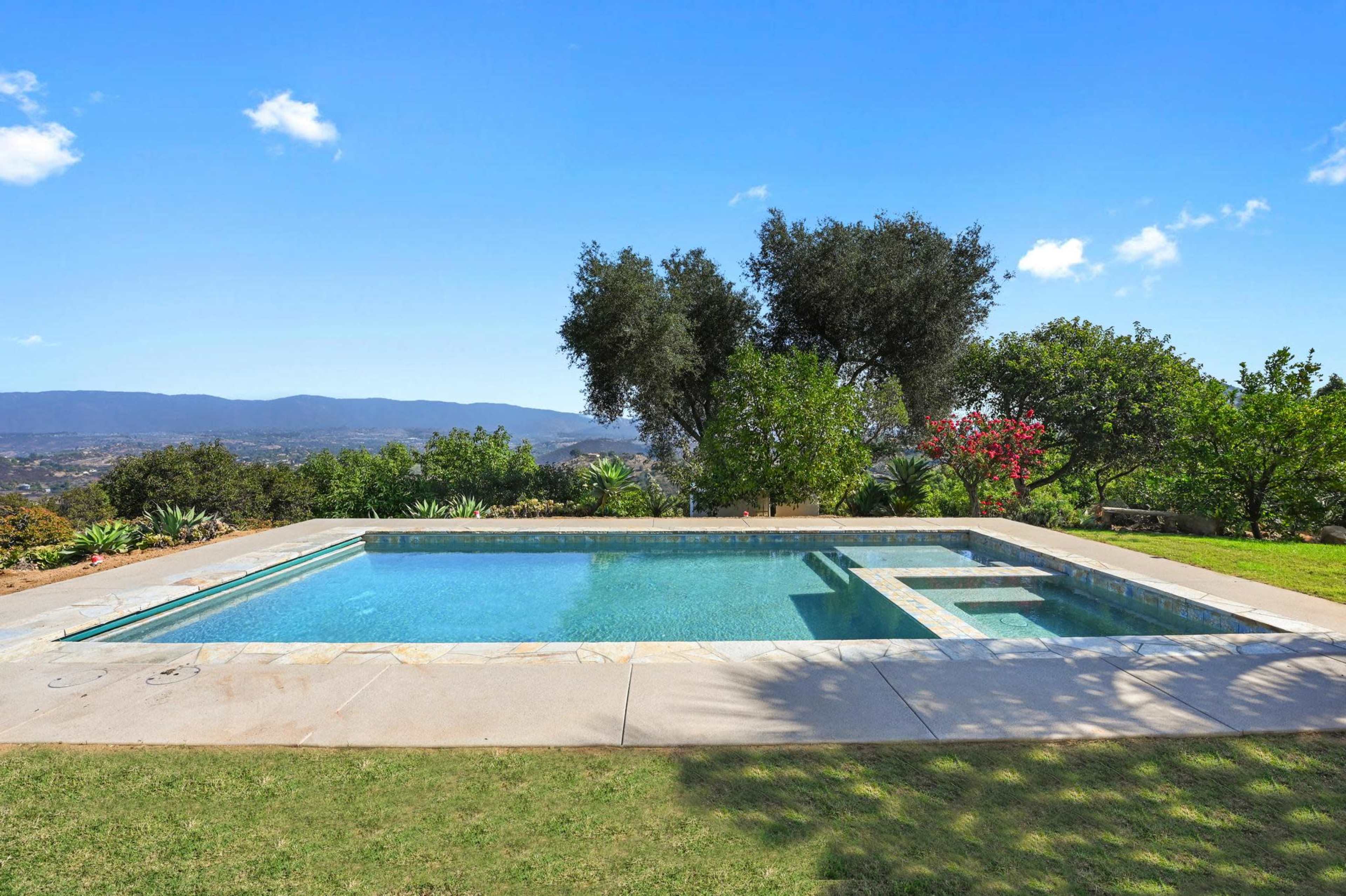 A rectangular swimming pool surrounded by grass and trees overlooks a mountainous landscape.