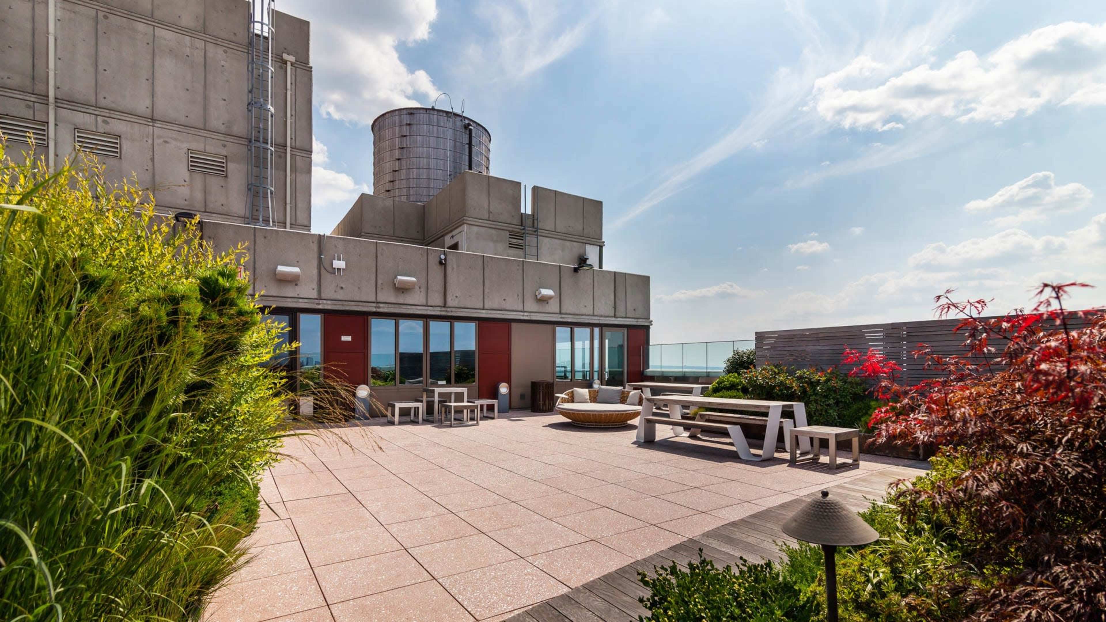 The image shows a rooftop terrace featuring a paved area with seating, surrounded by greenery and a large water tower in the background.