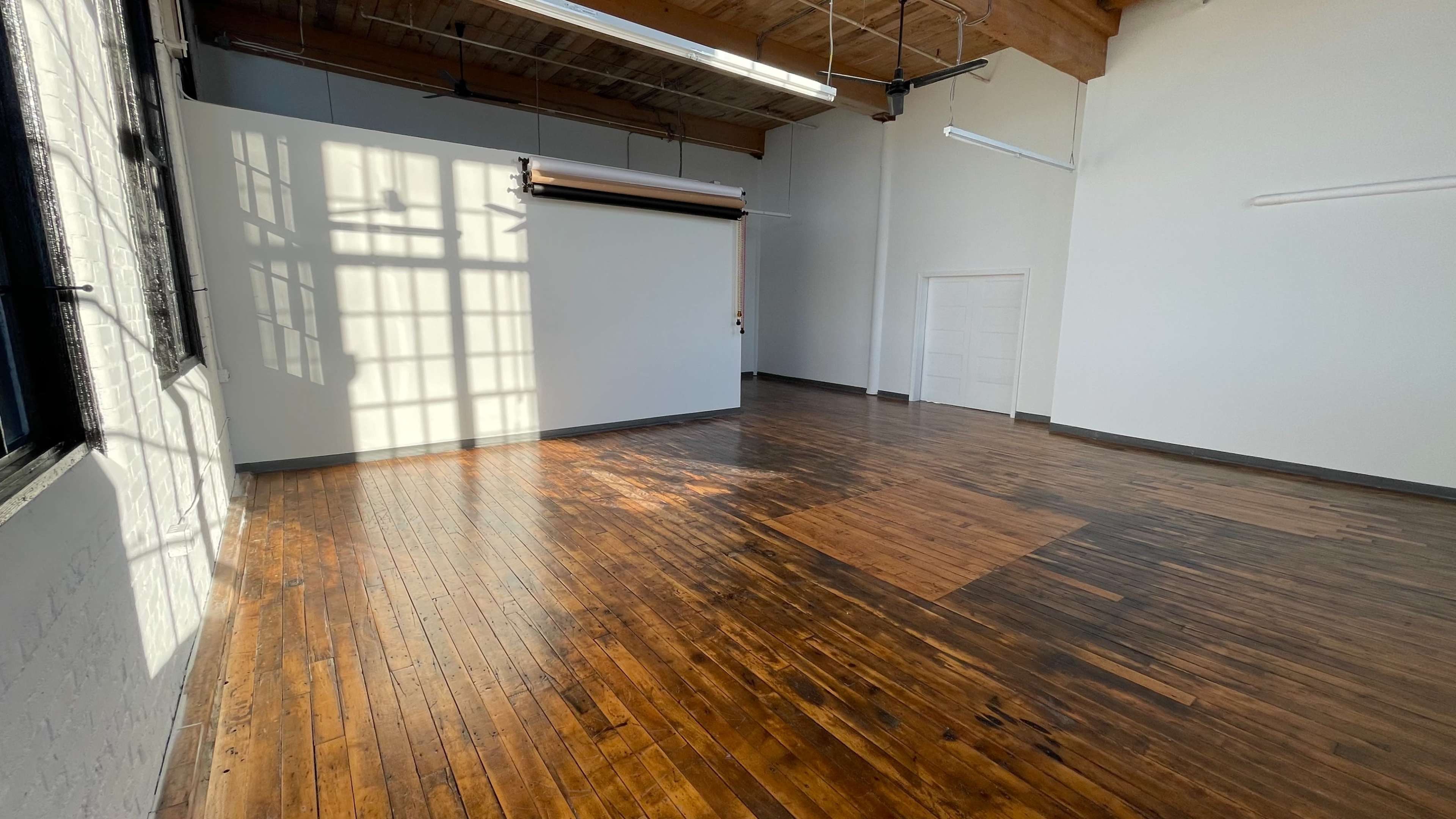 A spacious, well-lit room with wooden flooring, large windows, and a blank wall featuring a roll-up backdrop.