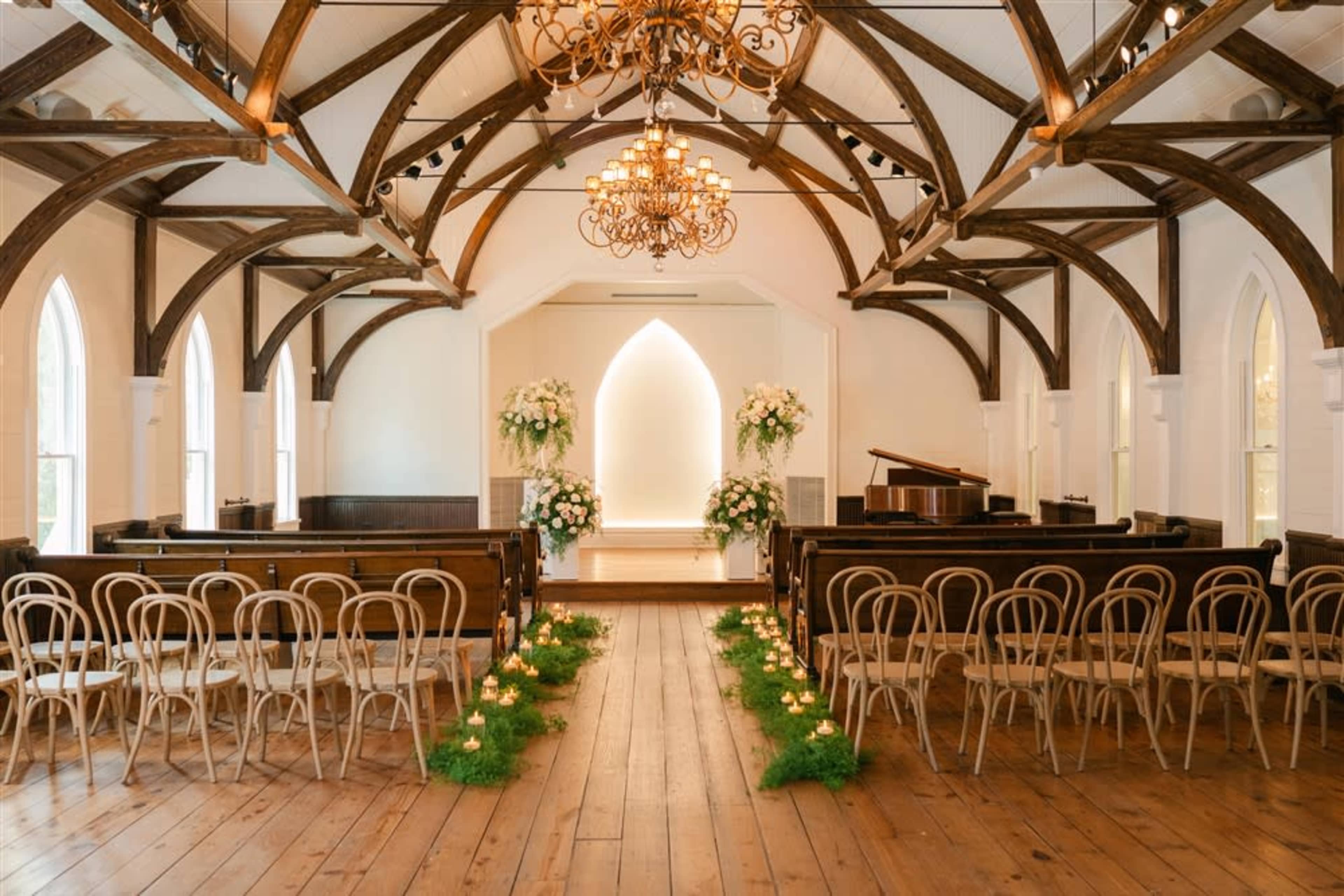 The image shows an elegantly arranged indoor ceremony space with wooden beams, benches facing an altar adorned with floral arrangements and candles lining the aisle.