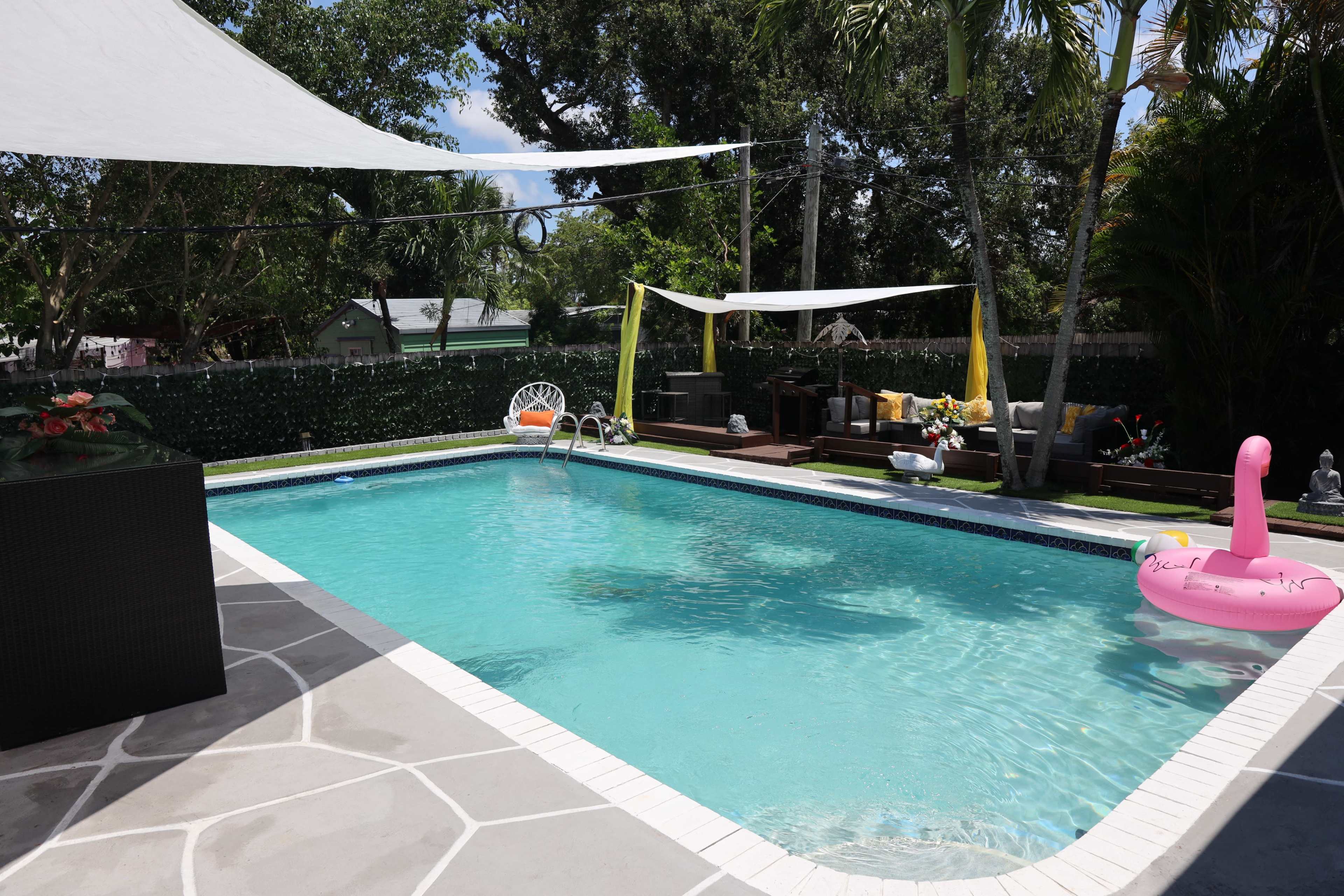 The image shows a backyard swimming pool surrounded by tropical plants, a pink inflatable flamingo, and lounge furniture partially shaded by fabric canopies.