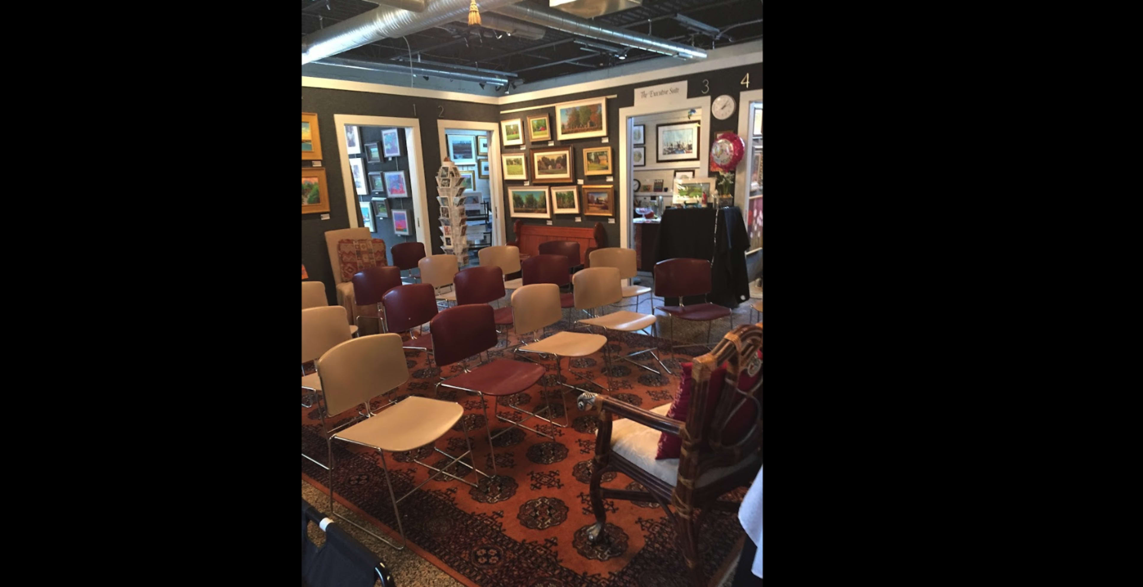 A gallery space features a seating arrangement of several chairs arranged in rows on an ornate rug, with artwork displayed on the walls.