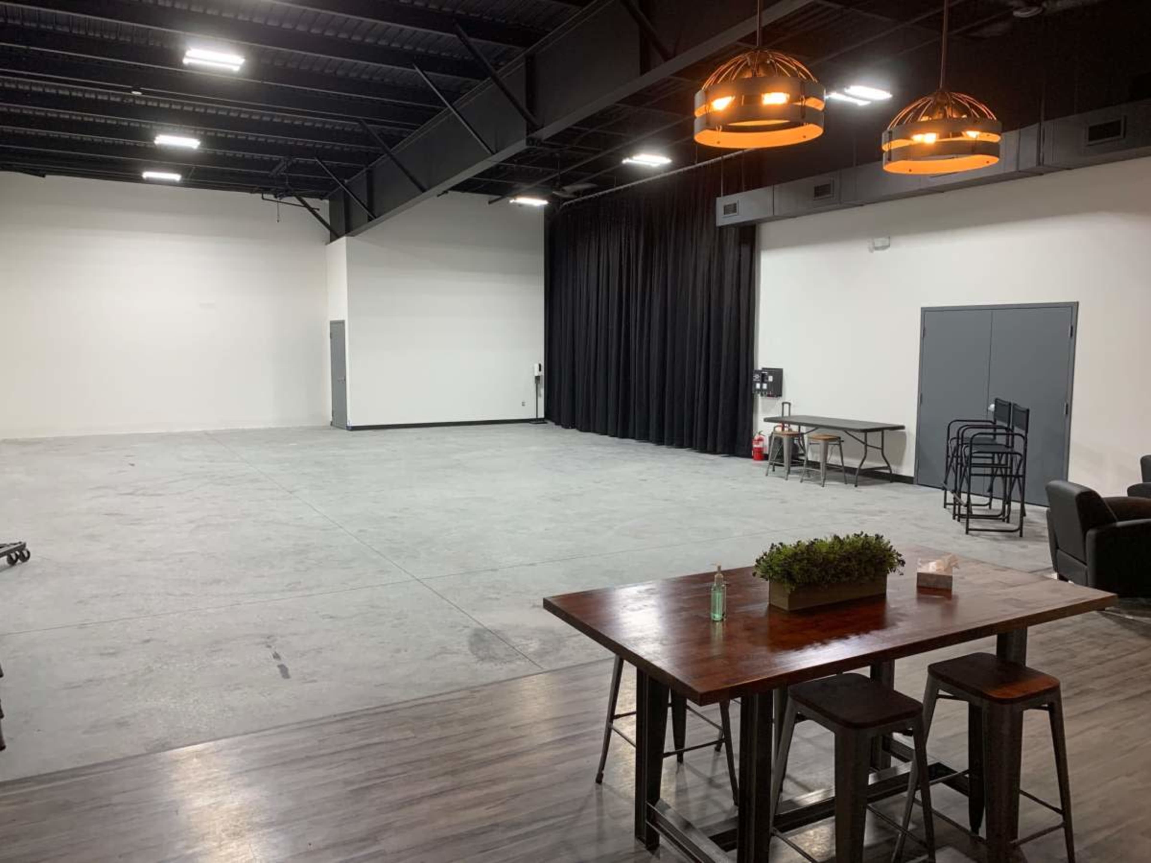 The image shows an empty industrial-style room with concrete flooring, a dark curtain on one wall, a wooden table and chairs, and modern pendant lights.