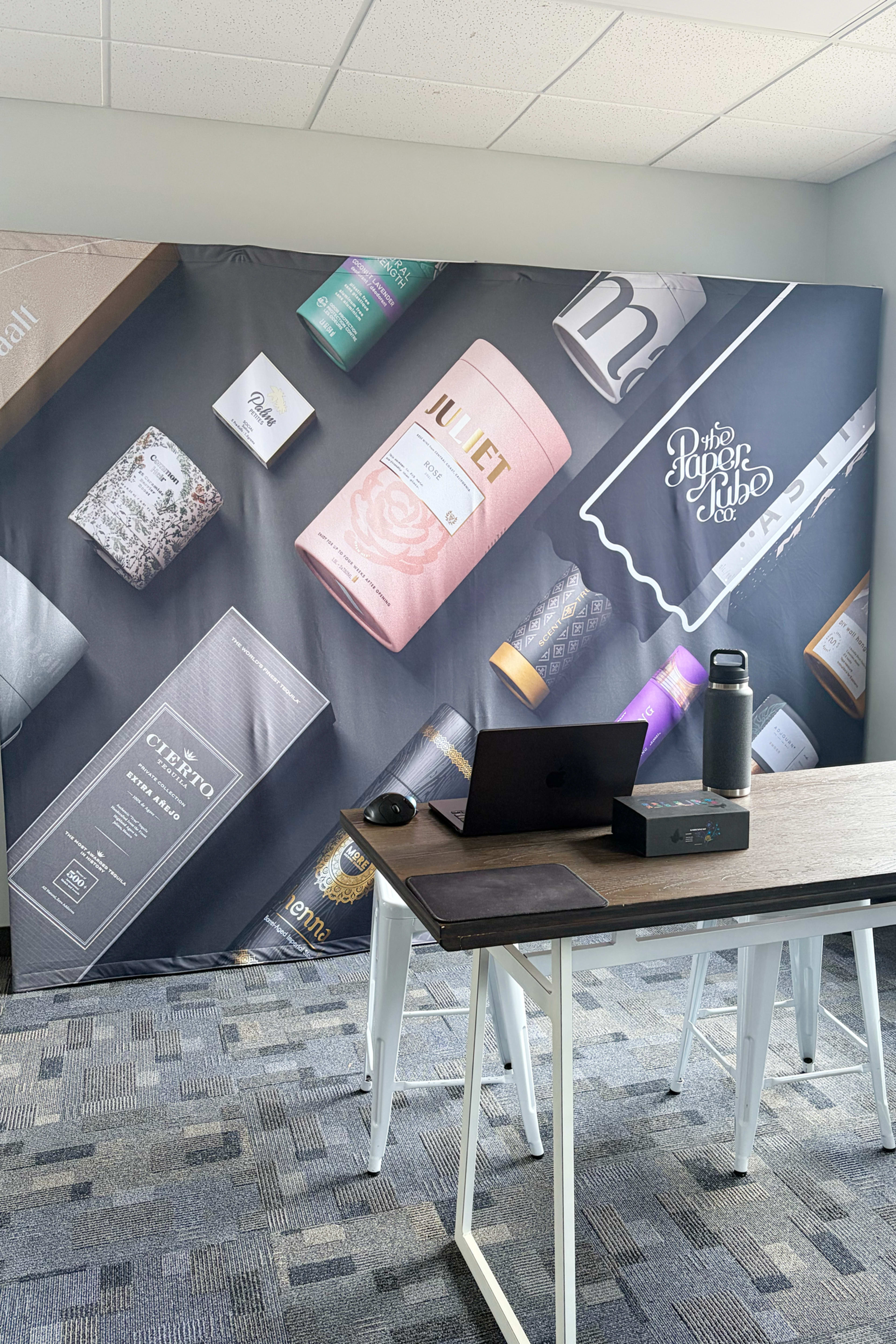 The image shows a workspace with a desk featuring a laptop, a water bottle, and a large backdrop displaying various product packages.