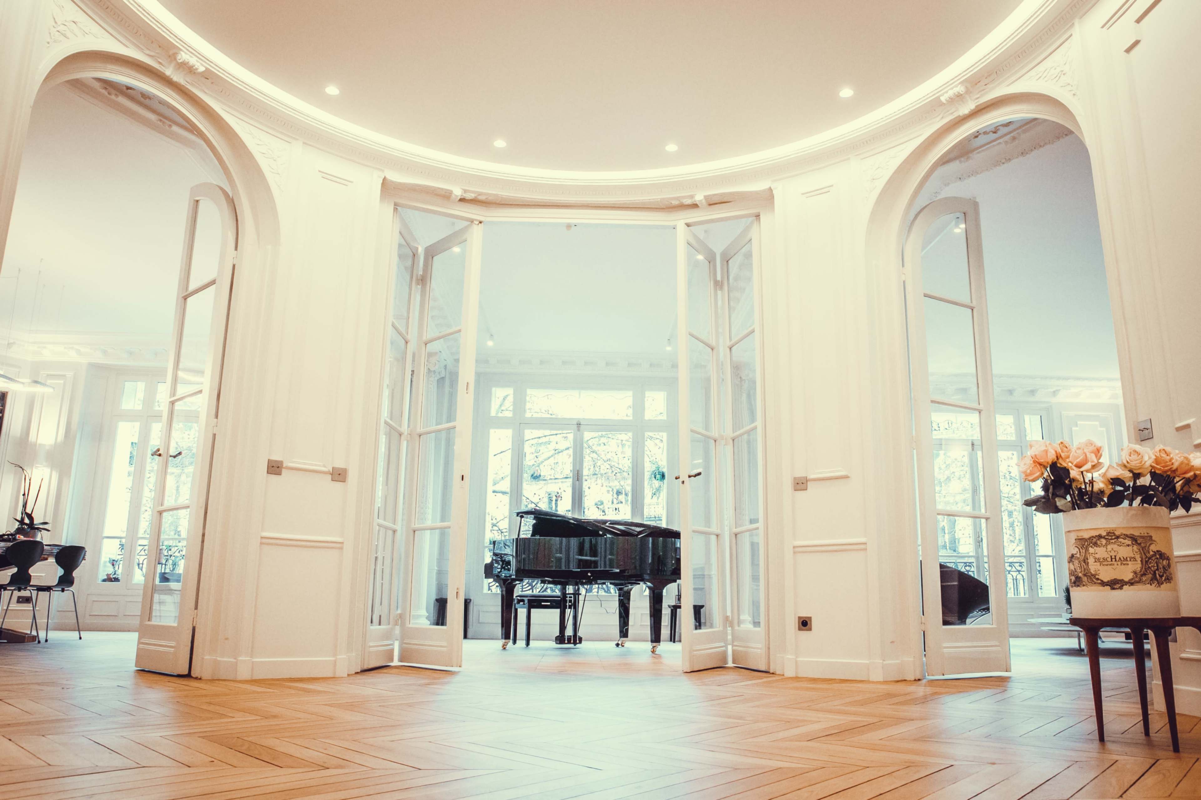 The image shows a spacious, elegantly designed room with large windows, featuring a grand piano at its center and white walls adorned with architectural details.