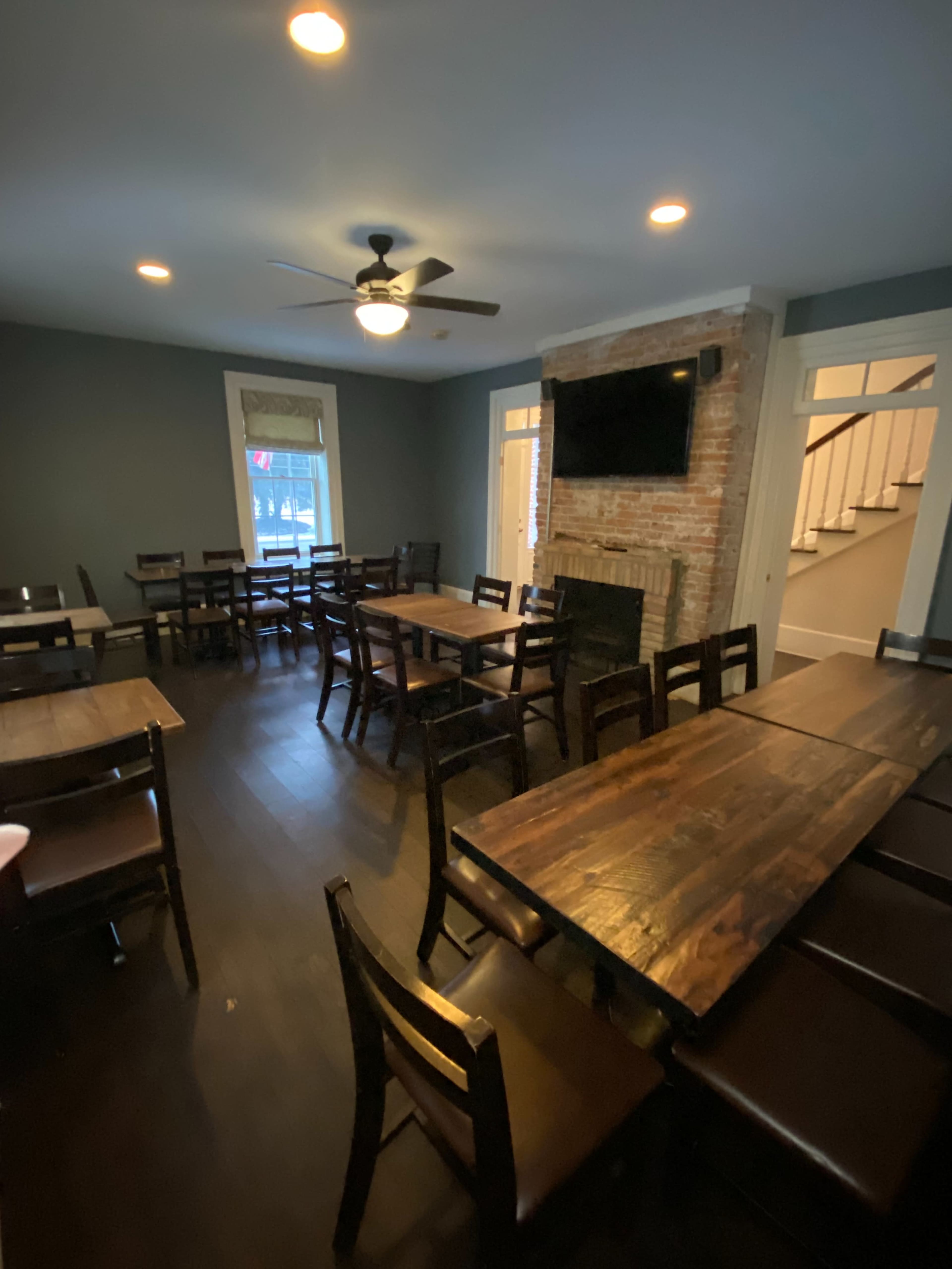 The image shows a dining room with wooden tables and chairs, featuring a brick wall and a television mounted on the wall, illuminated by overhead lights.