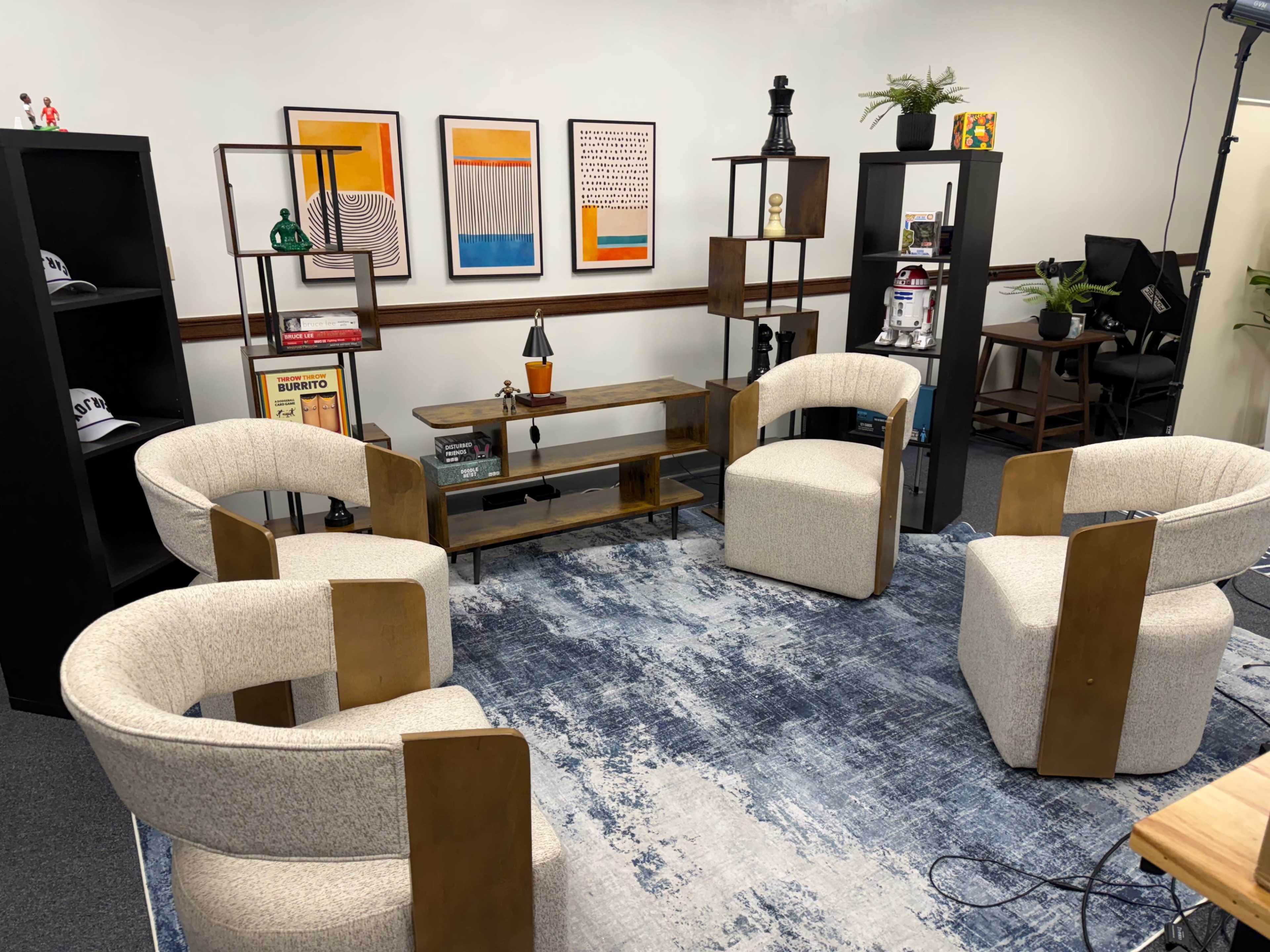 The image shows a modern office space featuring four cream-colored chairs arranged around a low wooden table, with shelves and framed artwork on the walls.
