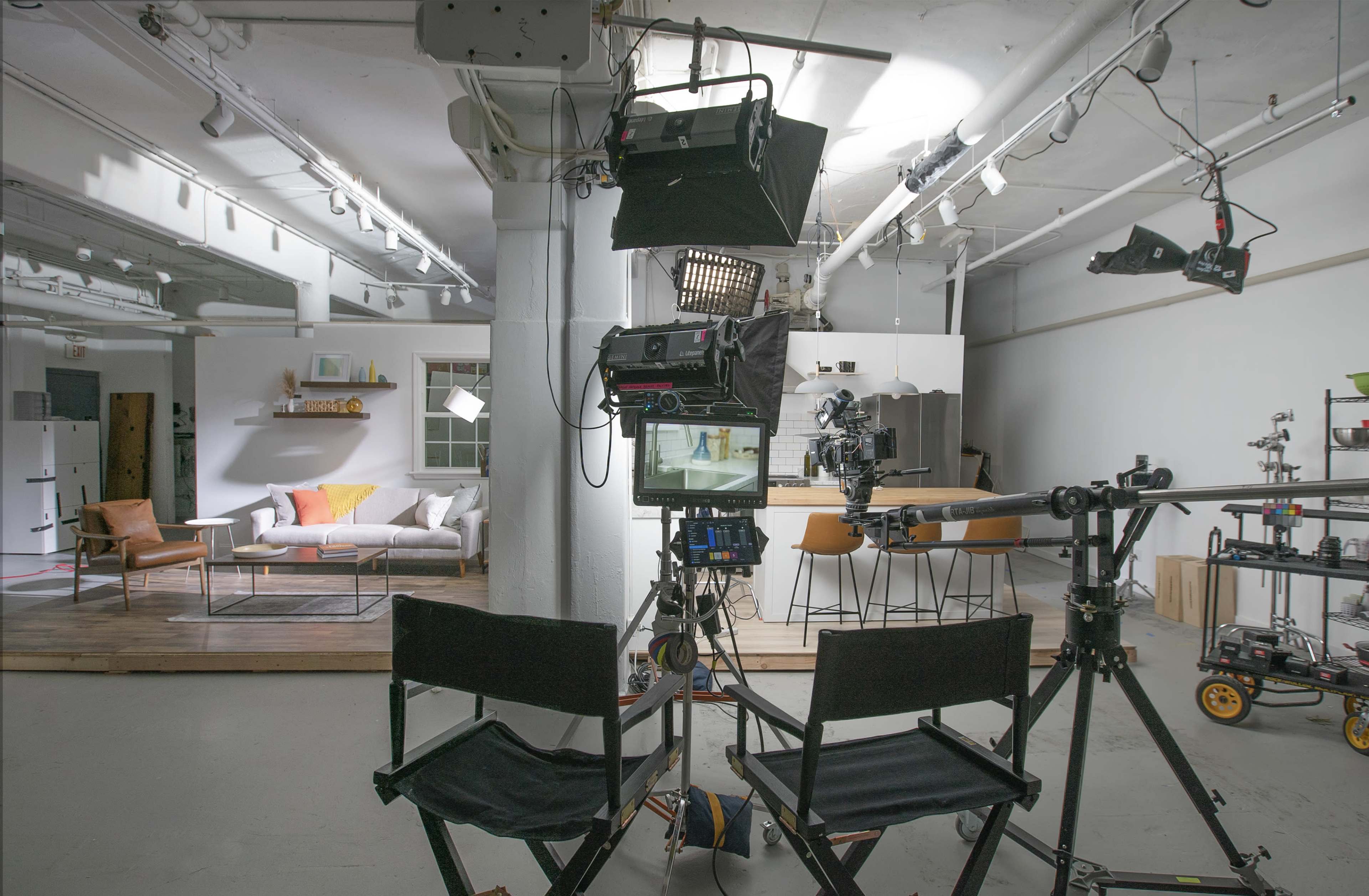 A television studio setup features two director's chairs facing a camera on a dolly, with lighting equipment above and a minimalistic living room in the background.