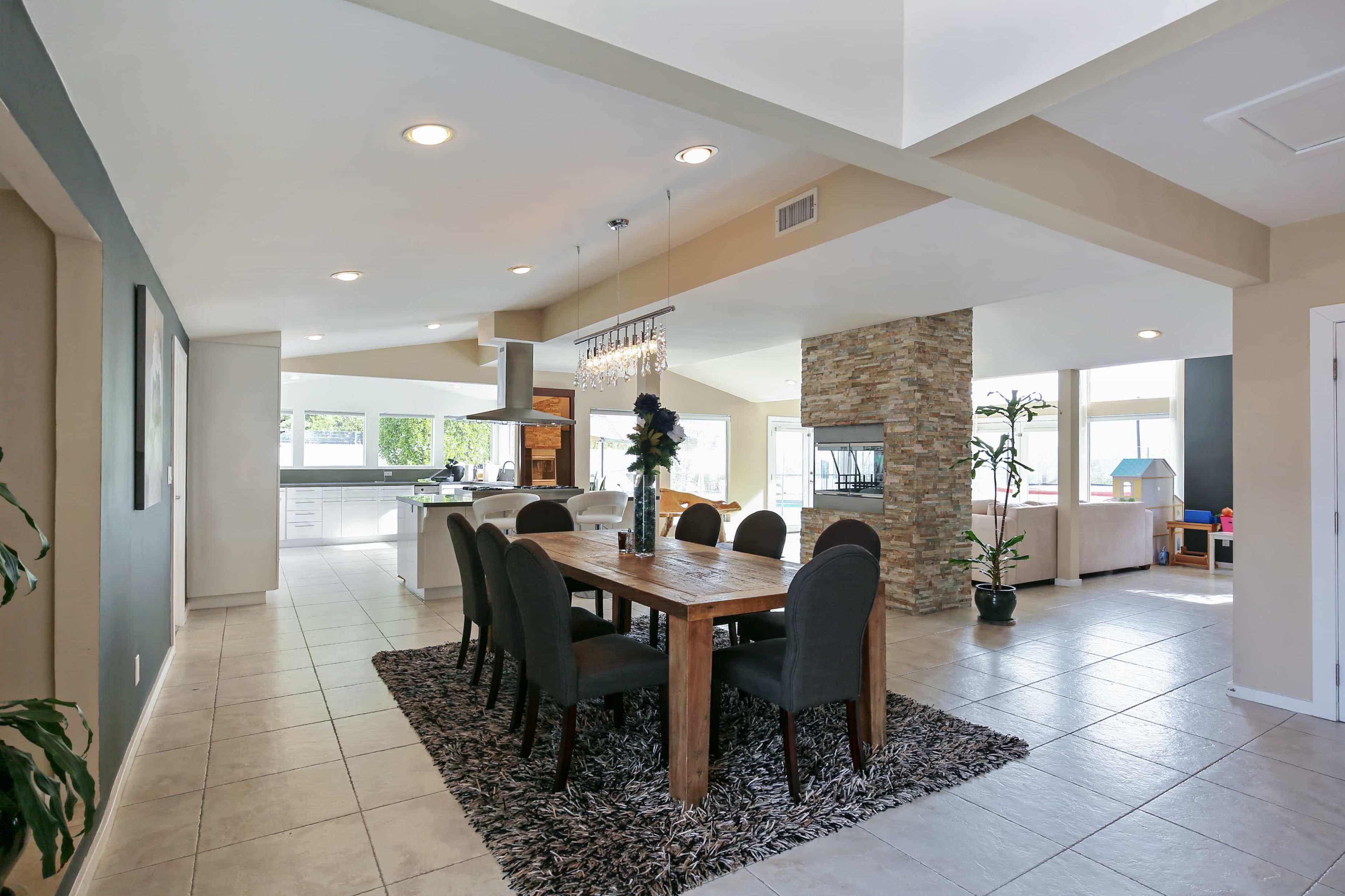 A modern dining area features a wooden table surrounded by chairs, set on a textured rug, with a stone fireplace and large windows in the background.