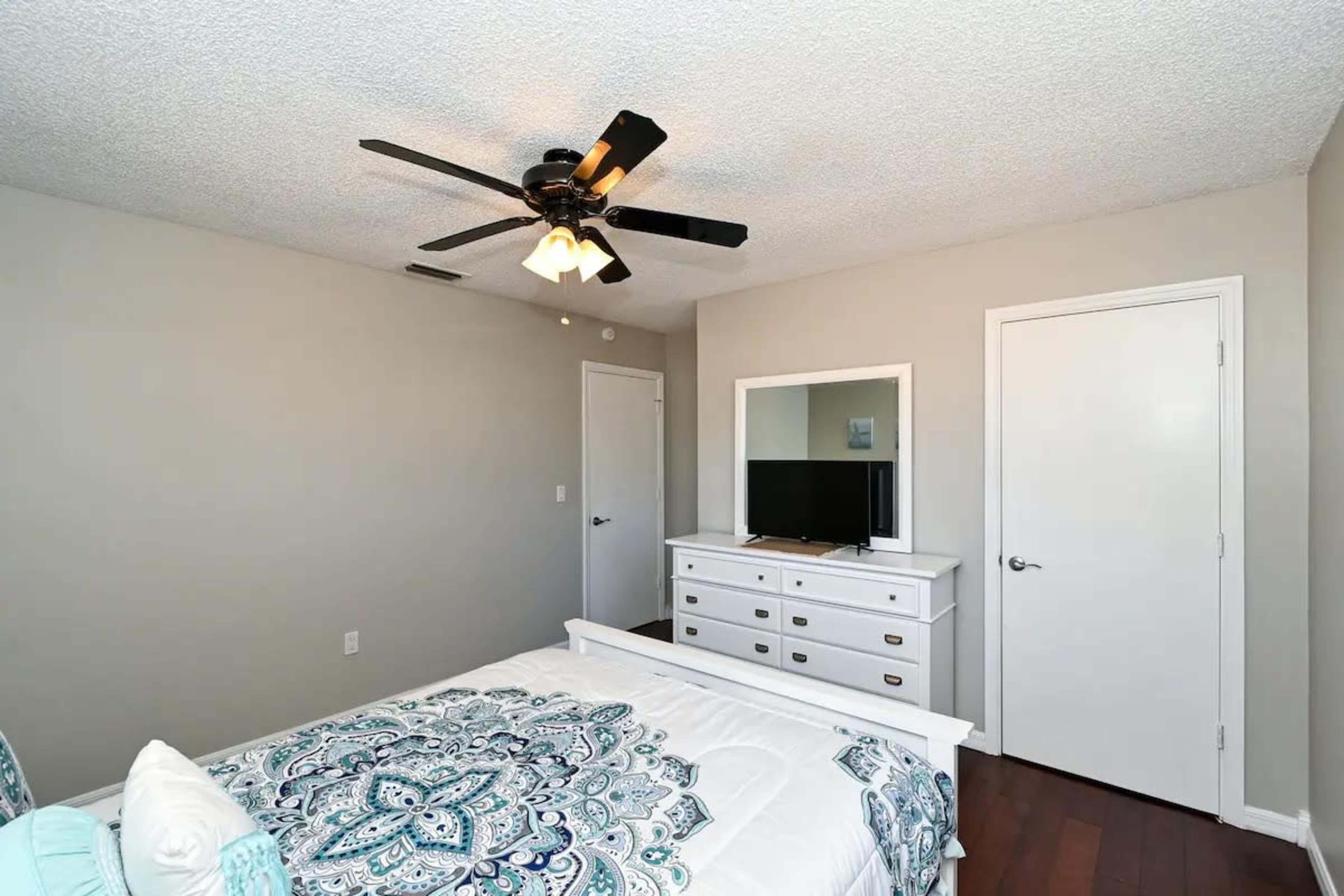 A simple bedroom features a bed with a patterned comforter, a dresser with a TV mounted on the wall, and a ceiling fan.