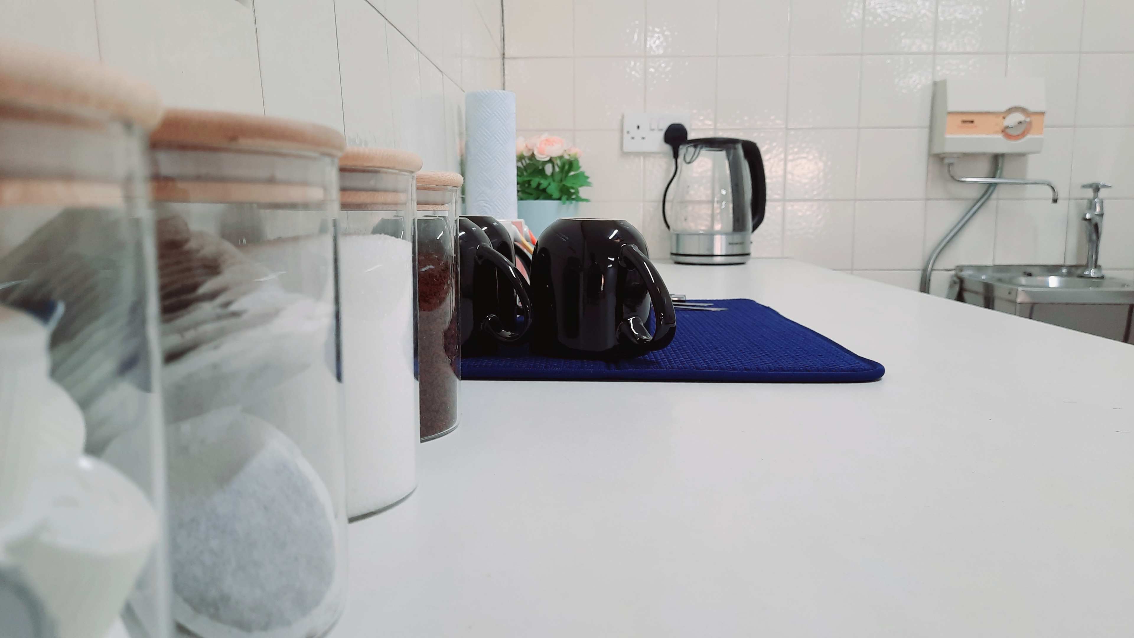 A kitchen countertop with glass containers, black coffee mugs, a kettle, and a roll of paper towels.