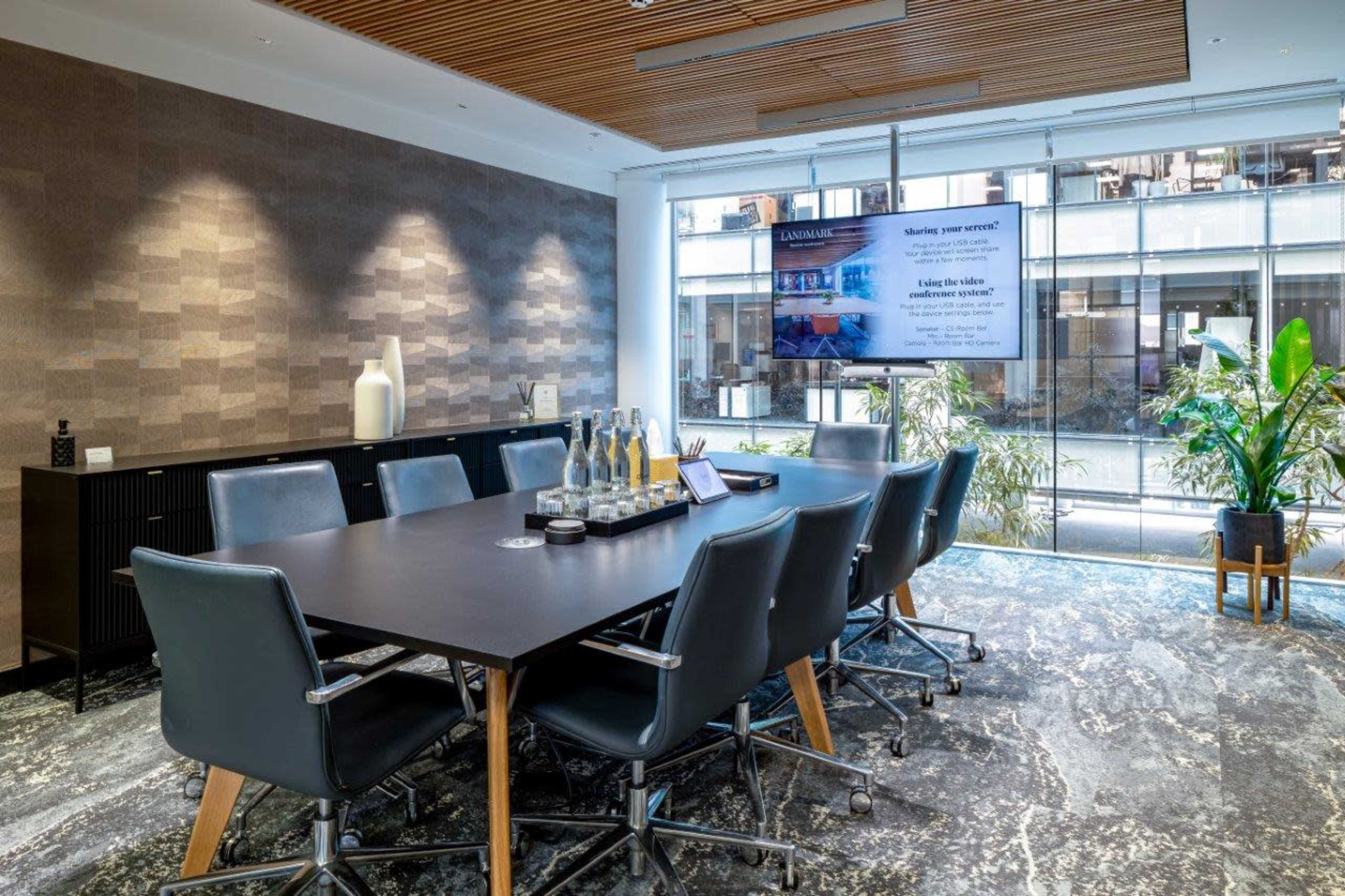 A modern conference room features a large table with ergonomic chairs, a television screen displaying a presentation, and plants visible through large windows.
