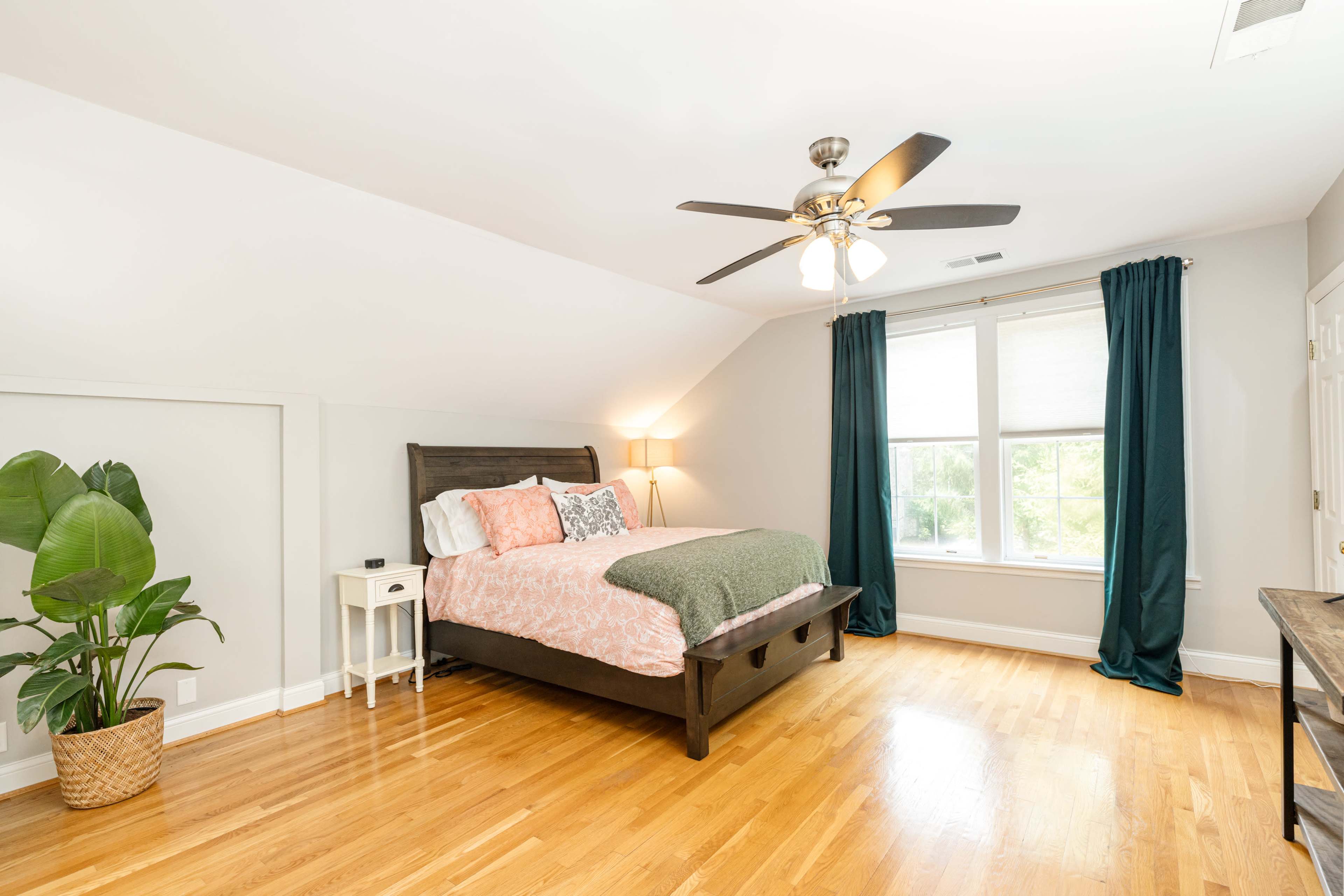 A well-lit bedroom featuring a bed with a green and pink comforter, a ceiling fan, large windows with curtains, and a bedside table with a lamp next to a potted plant.