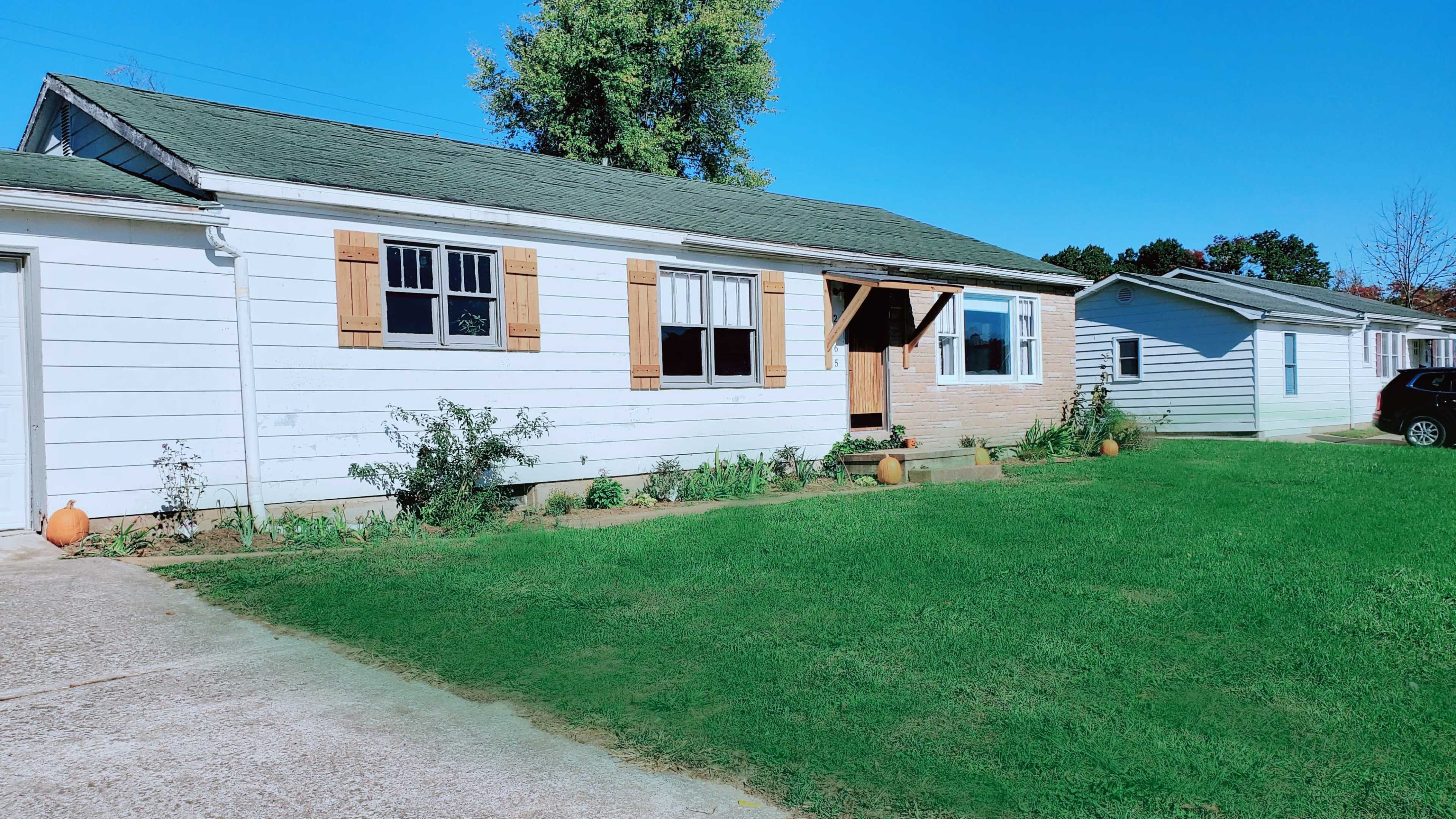 A single-story house with white siding and wooden shutters sits on a grassy lawn alongside a concrete driveway.