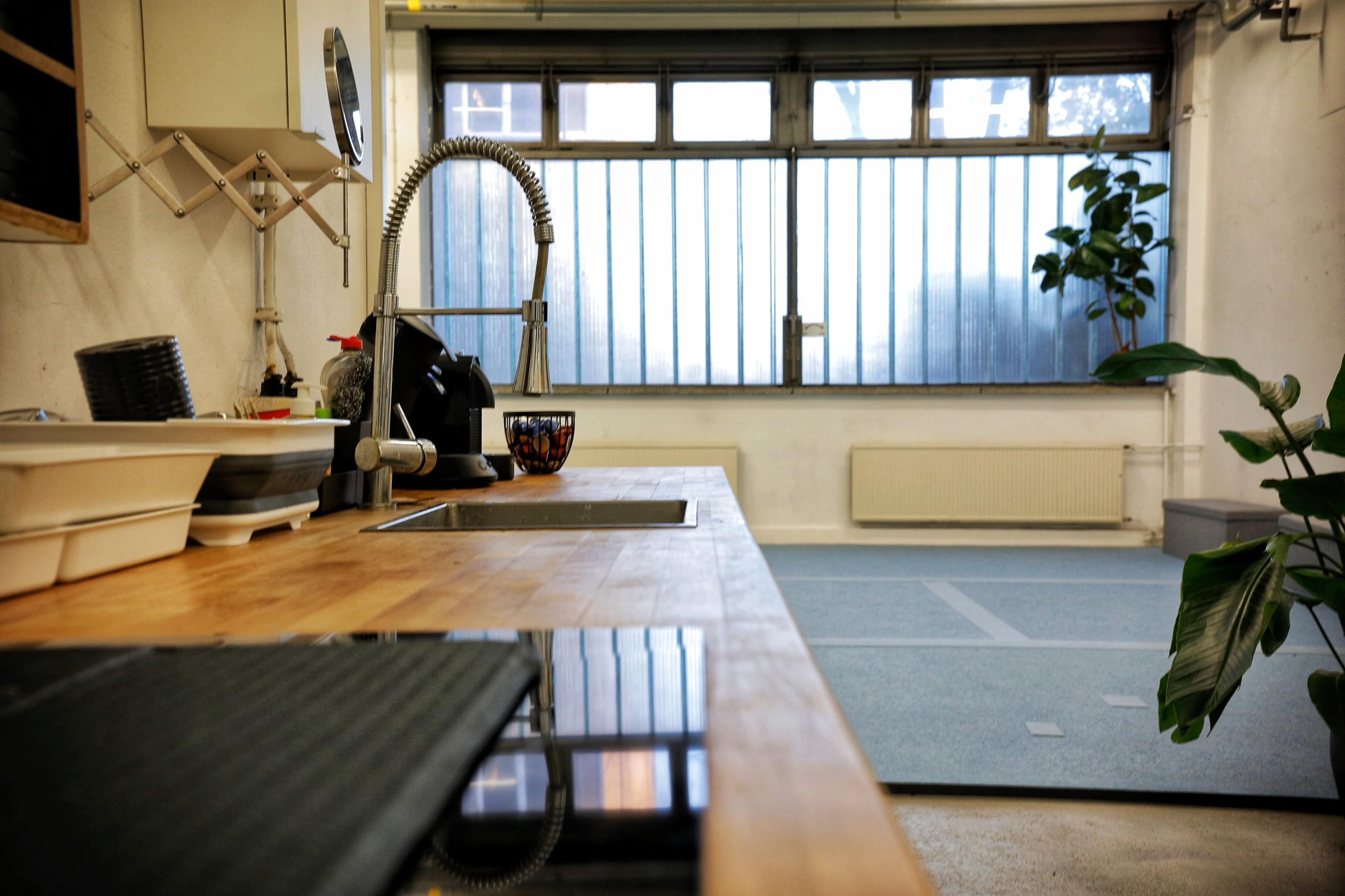 The image shows a modern kitchen with a wooden countertop, a sink, an induction cooktop, and a large window allowing natural light to enter the room.