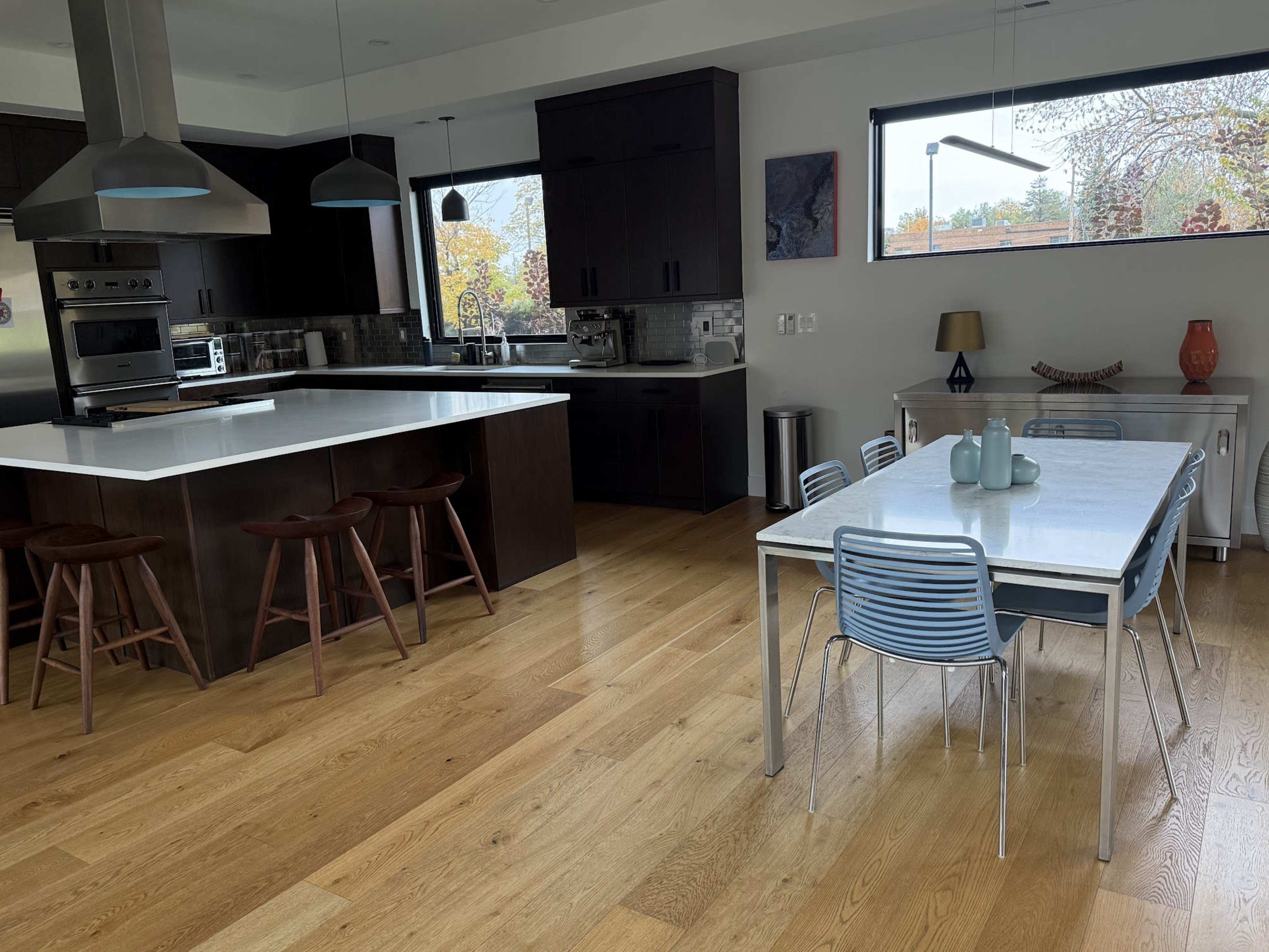 The image shows a modern kitchen with dark cabinetry, a central island with bar stools, and a dining area equipped with a metal table and chairs.