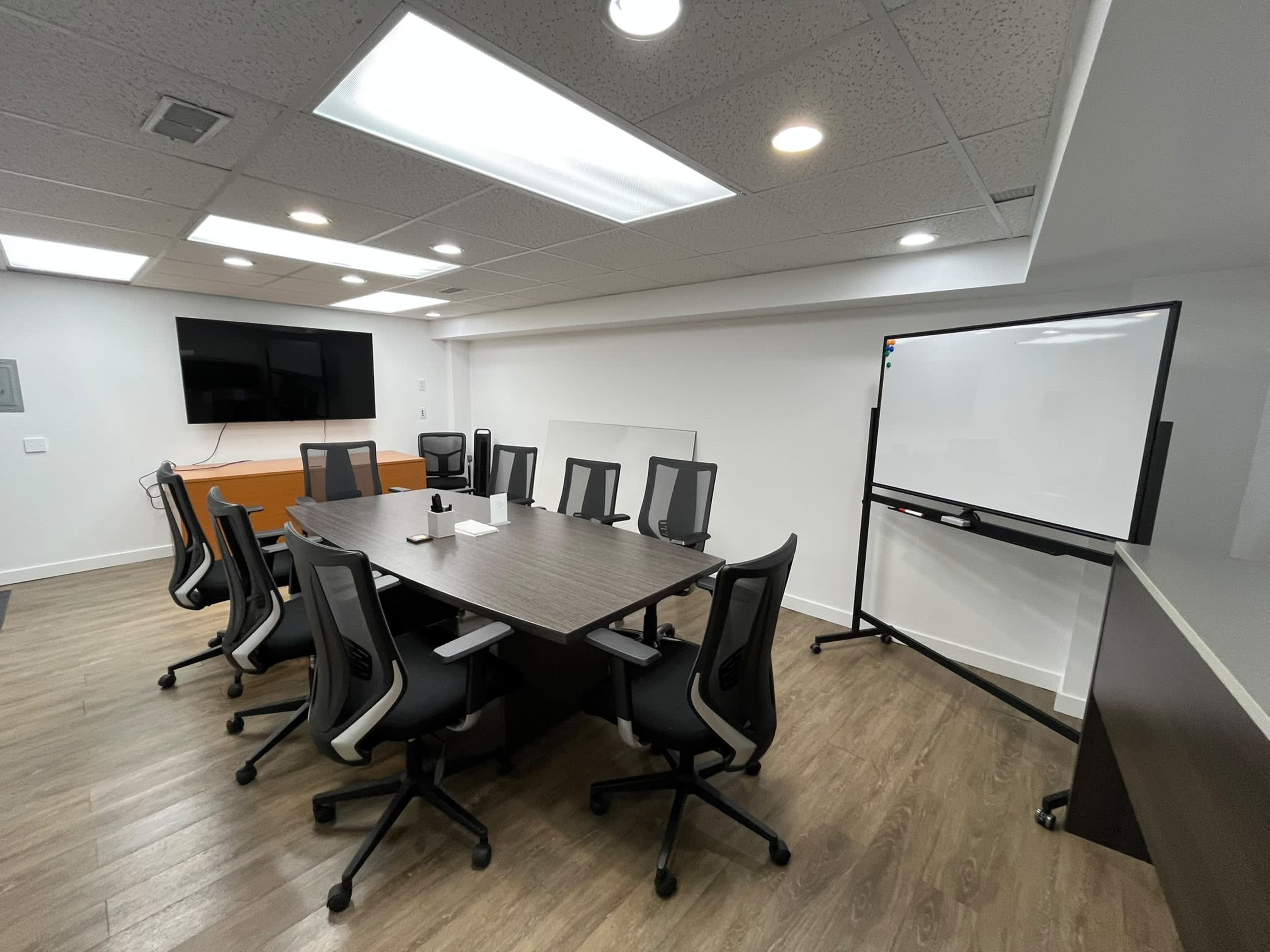 A modern conference room is equipped with a long table, ergonomic chairs, a large screen mounted on the wall, and a whiteboard on a mobile stand.