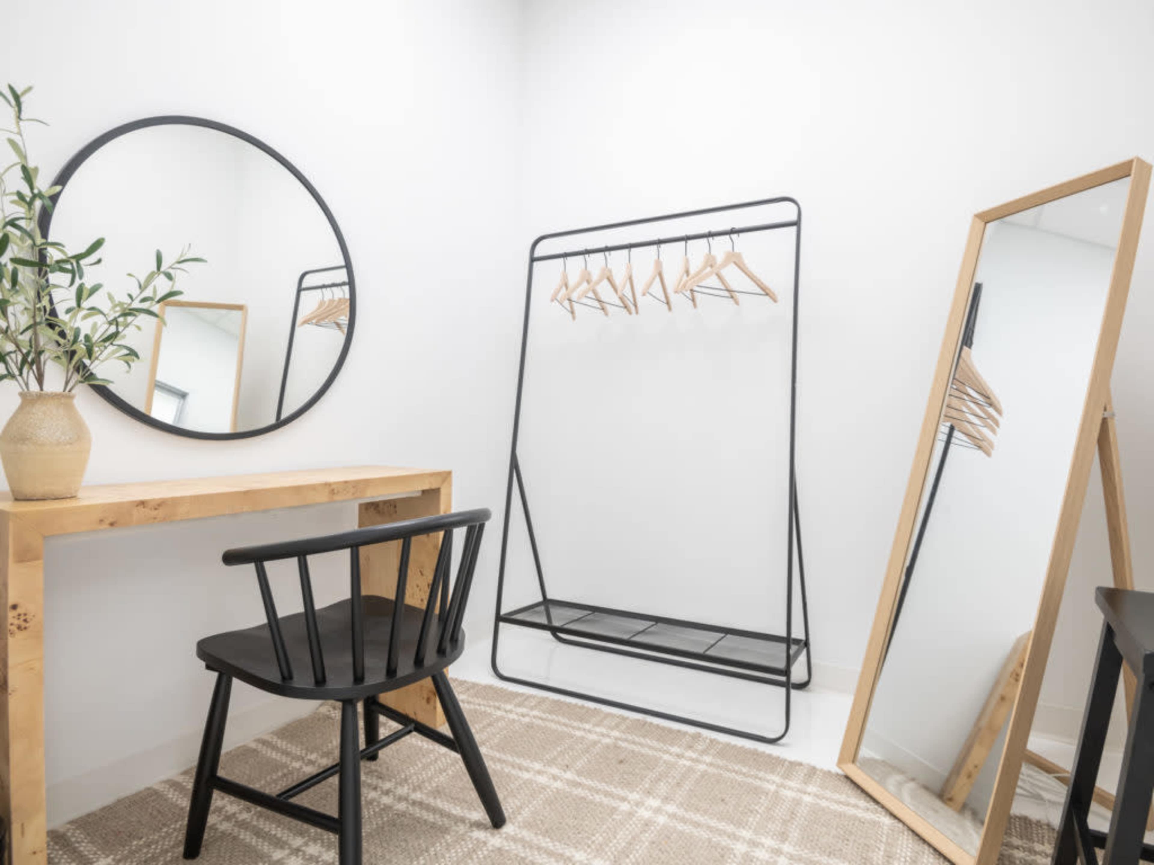 A minimalist dressing area featuring a round mirror, a wooden desk with a black chair, a clothing rack with hangers, and a standing mirror against a white wall.