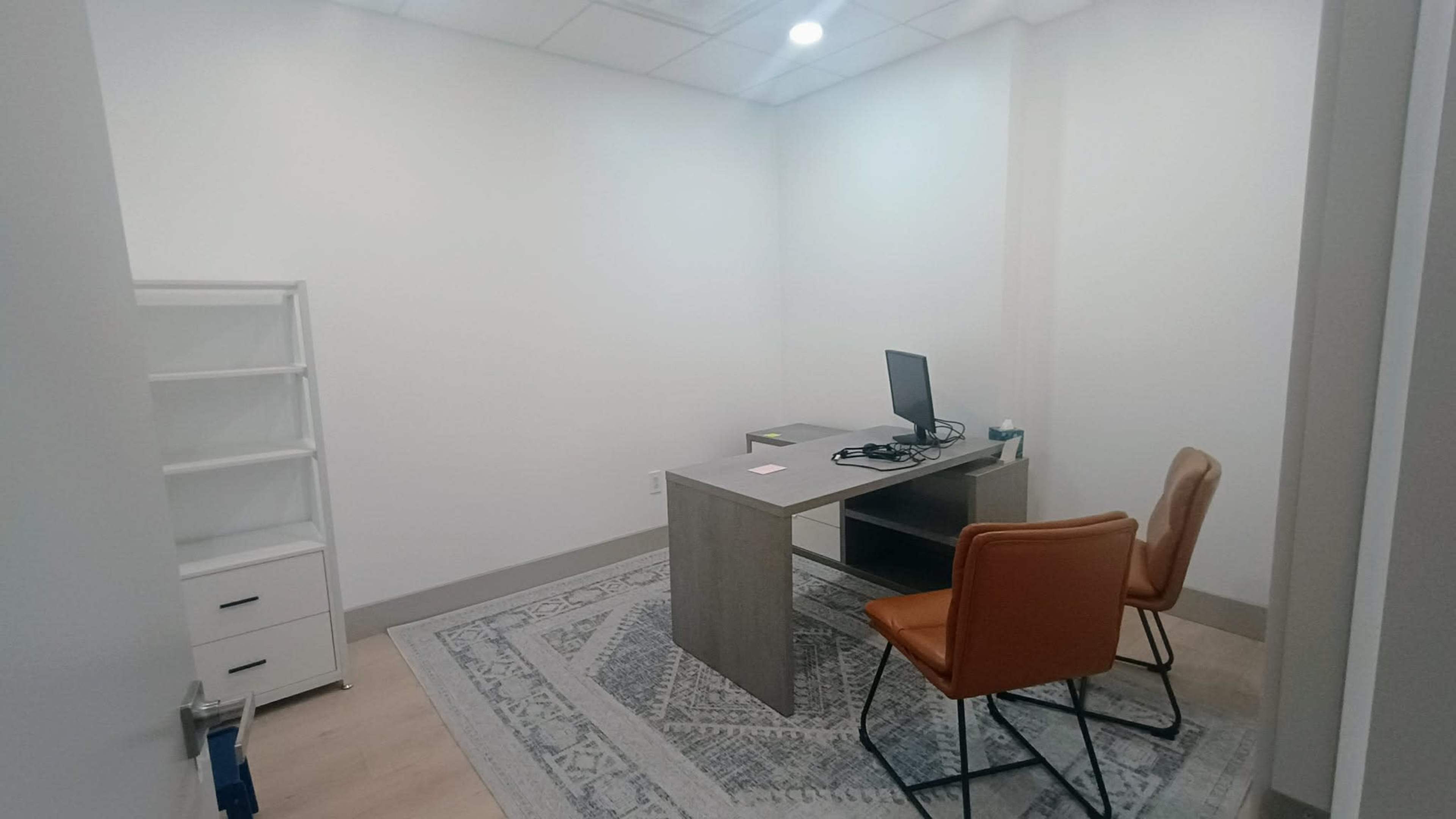 The image shows a minimalist office space with a desk, a computer, two chairs, and a shelf against a white wall.