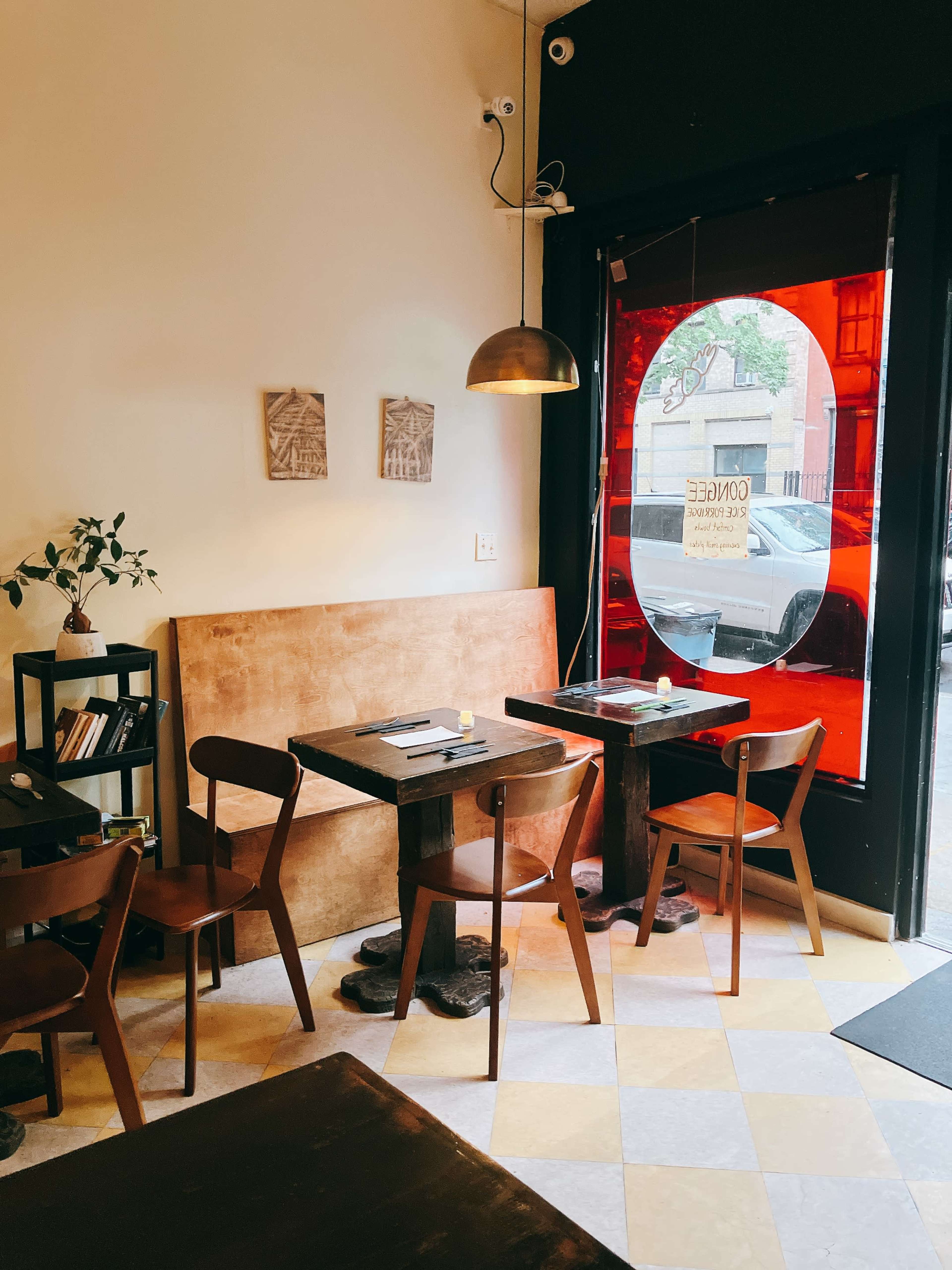 The image shows a small café interior with wooden tables and chairs, a potted plant on a shelf, and a large round red window.