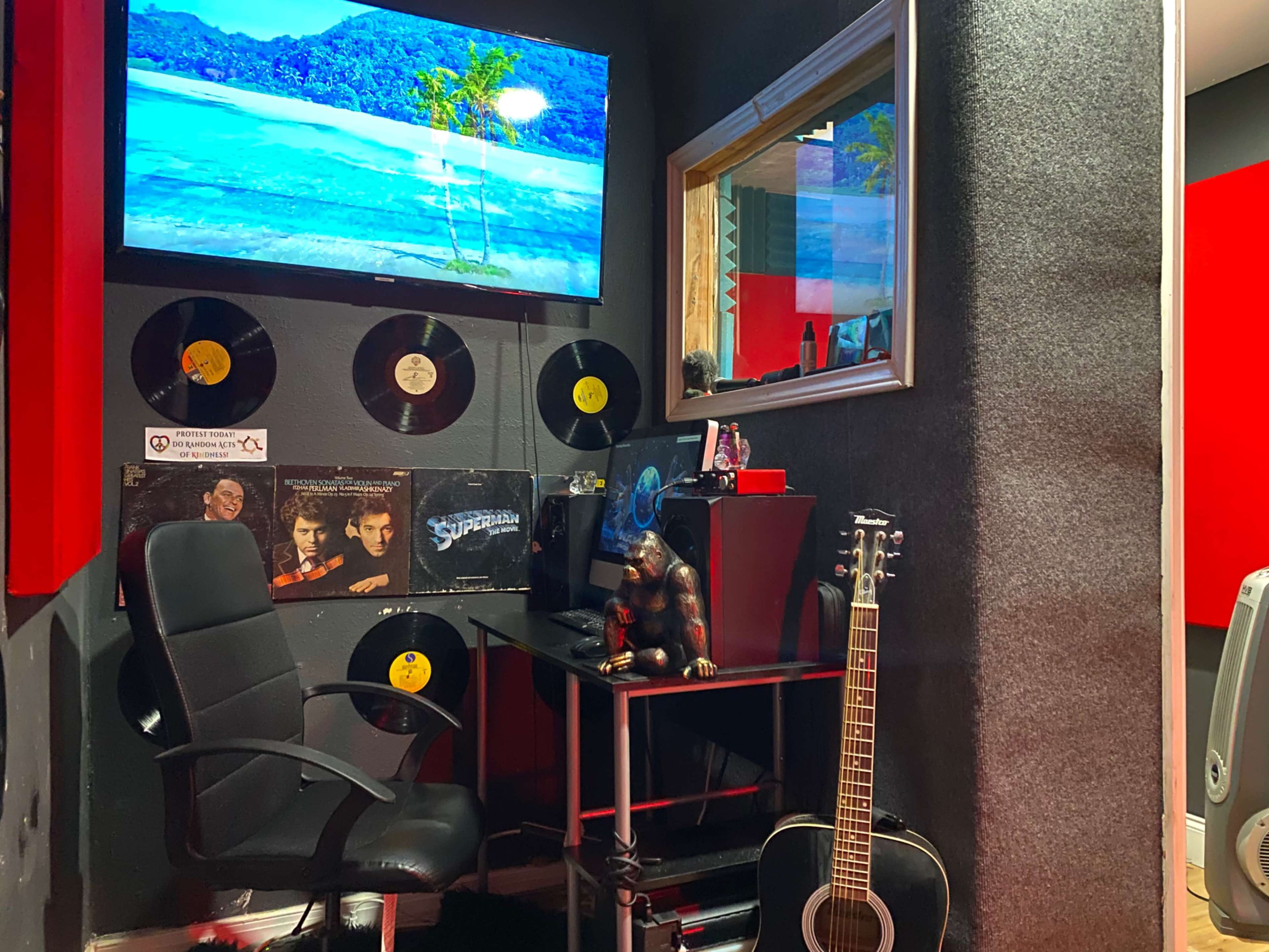 A music studio corner with a black chair, a desk holding a computer and a sculpture, a guitar leaning against the wall, and records displayed alongside a TV screen showing a beach scene.