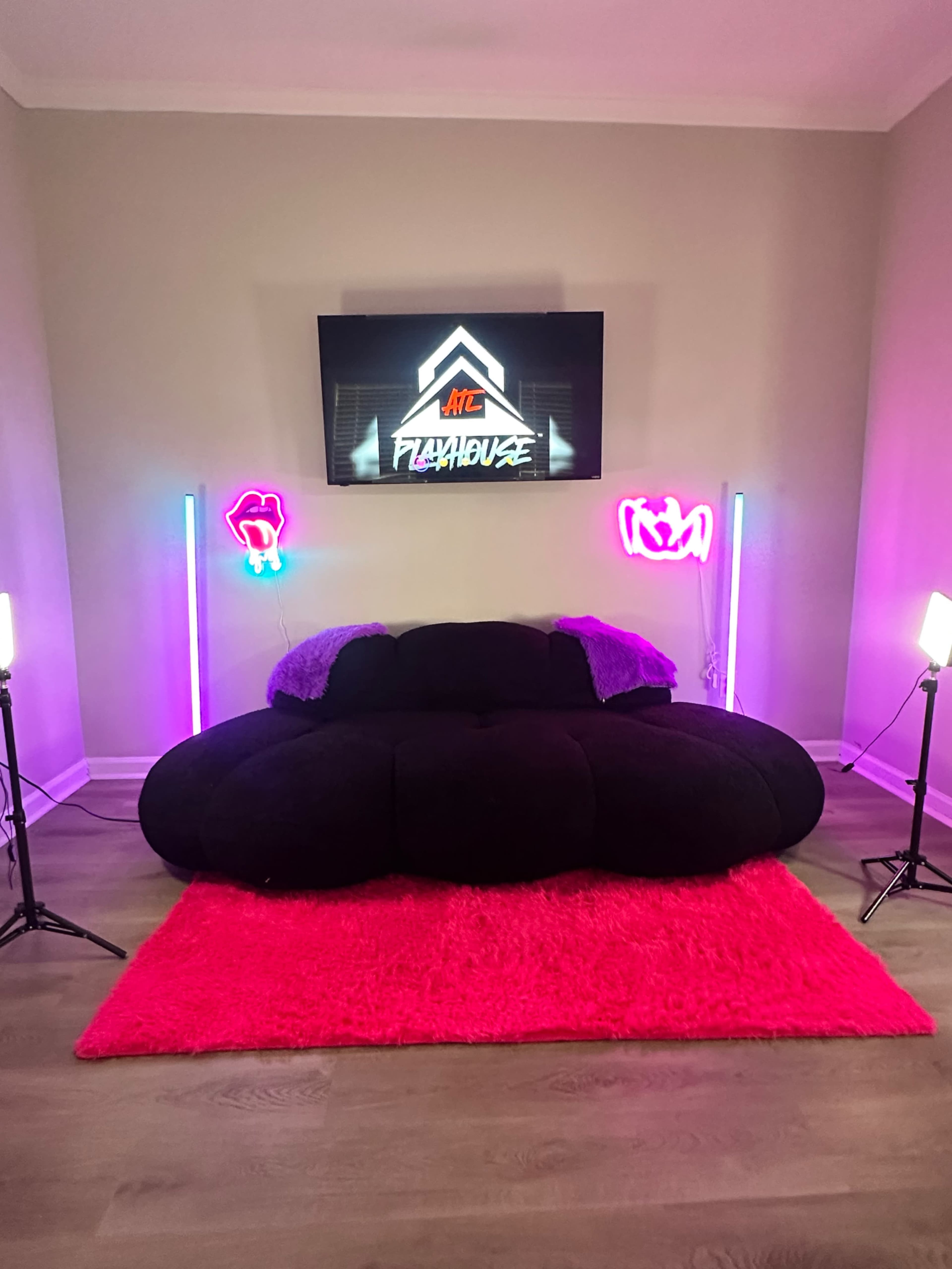 The image shows a black, cloud-shaped couch on a bright pink rug, with neon lights and a television displaying the words "Playhouse" in the background.