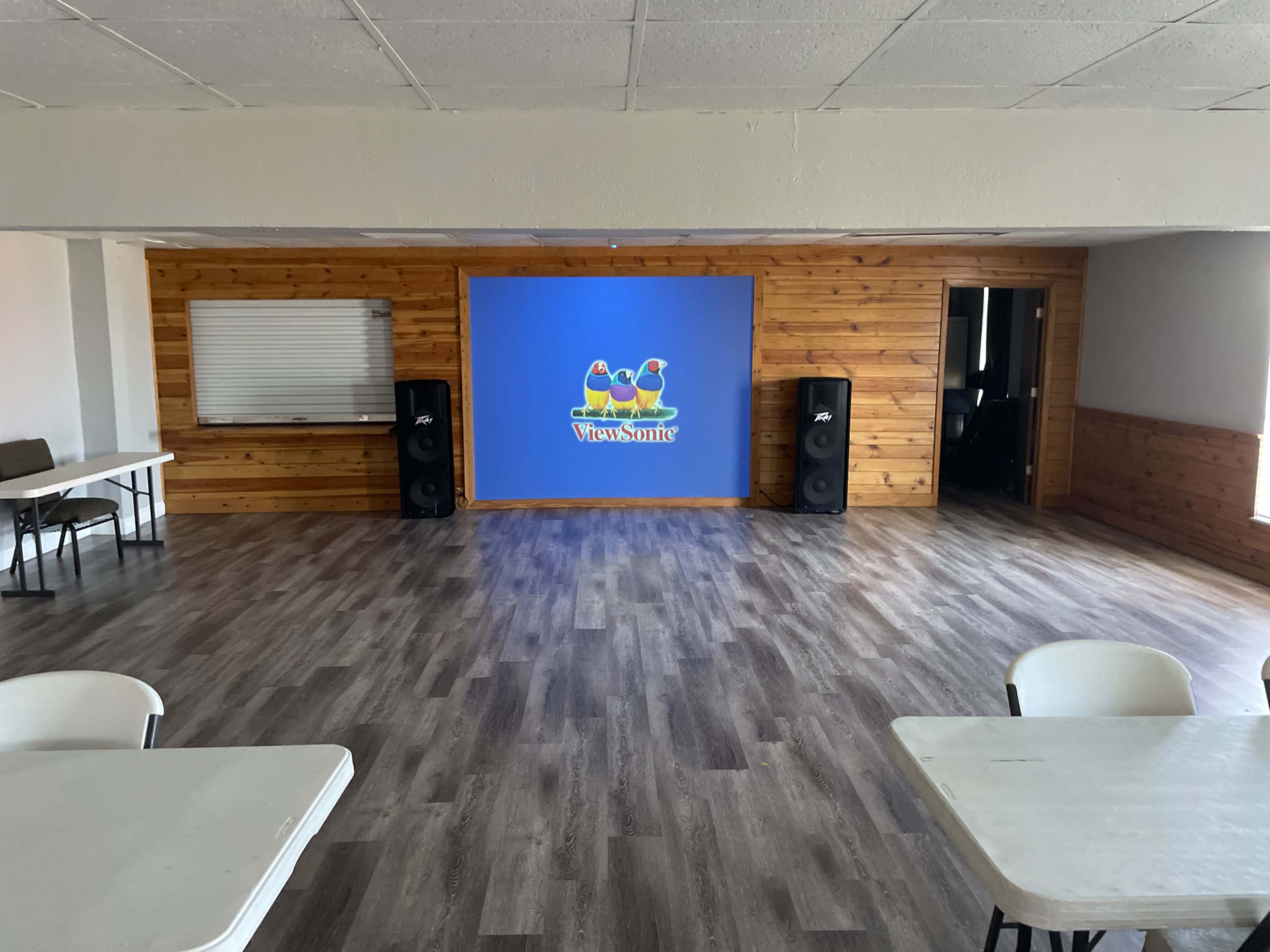 The image shows a spacious, empty room with wooden walls, two large speakers on either side of a blue display screen, and several white tables arranged throughout.
