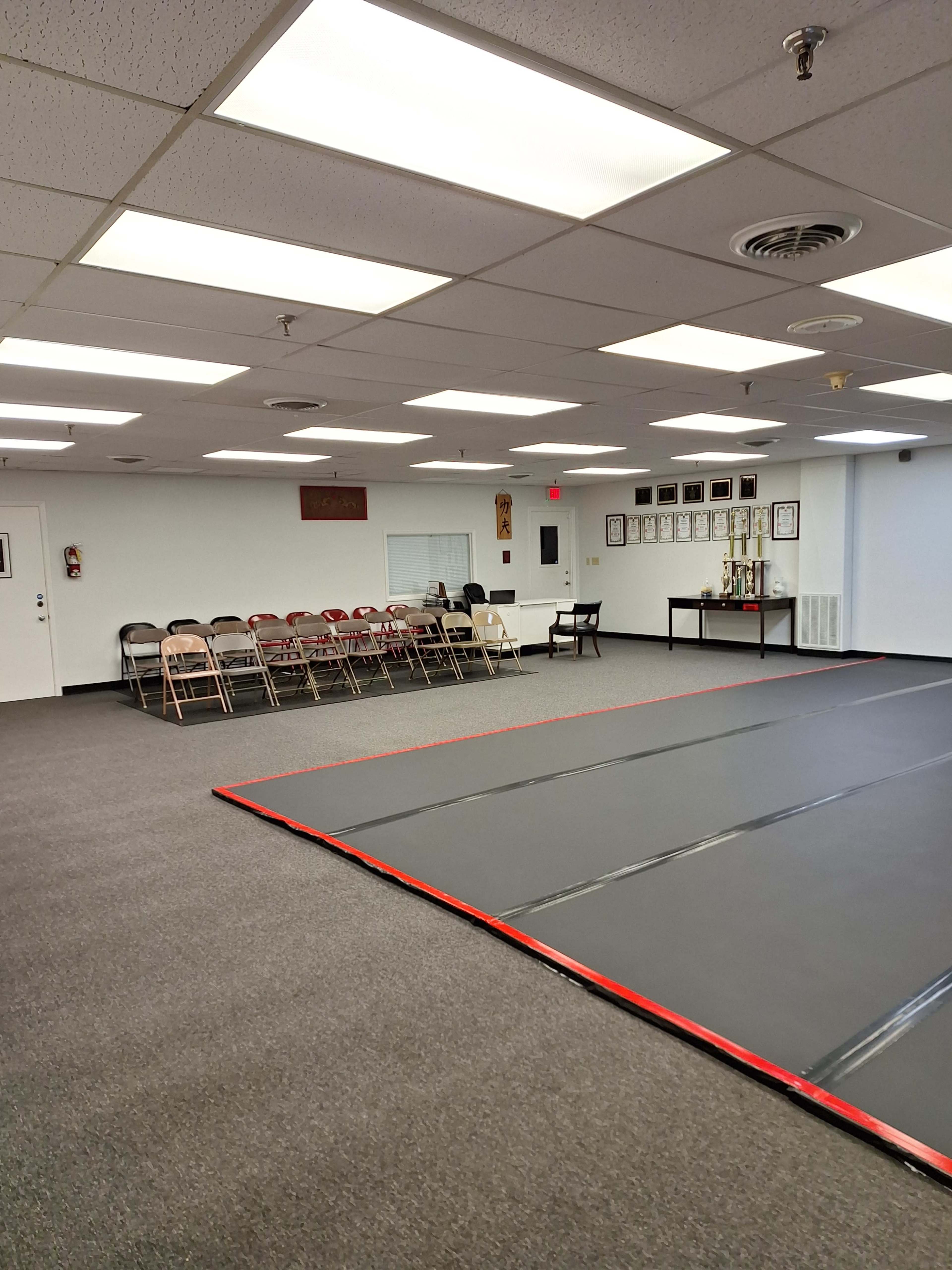 The image shows a spacious martial arts studio with a mat area and rows of folding chairs along one wall.