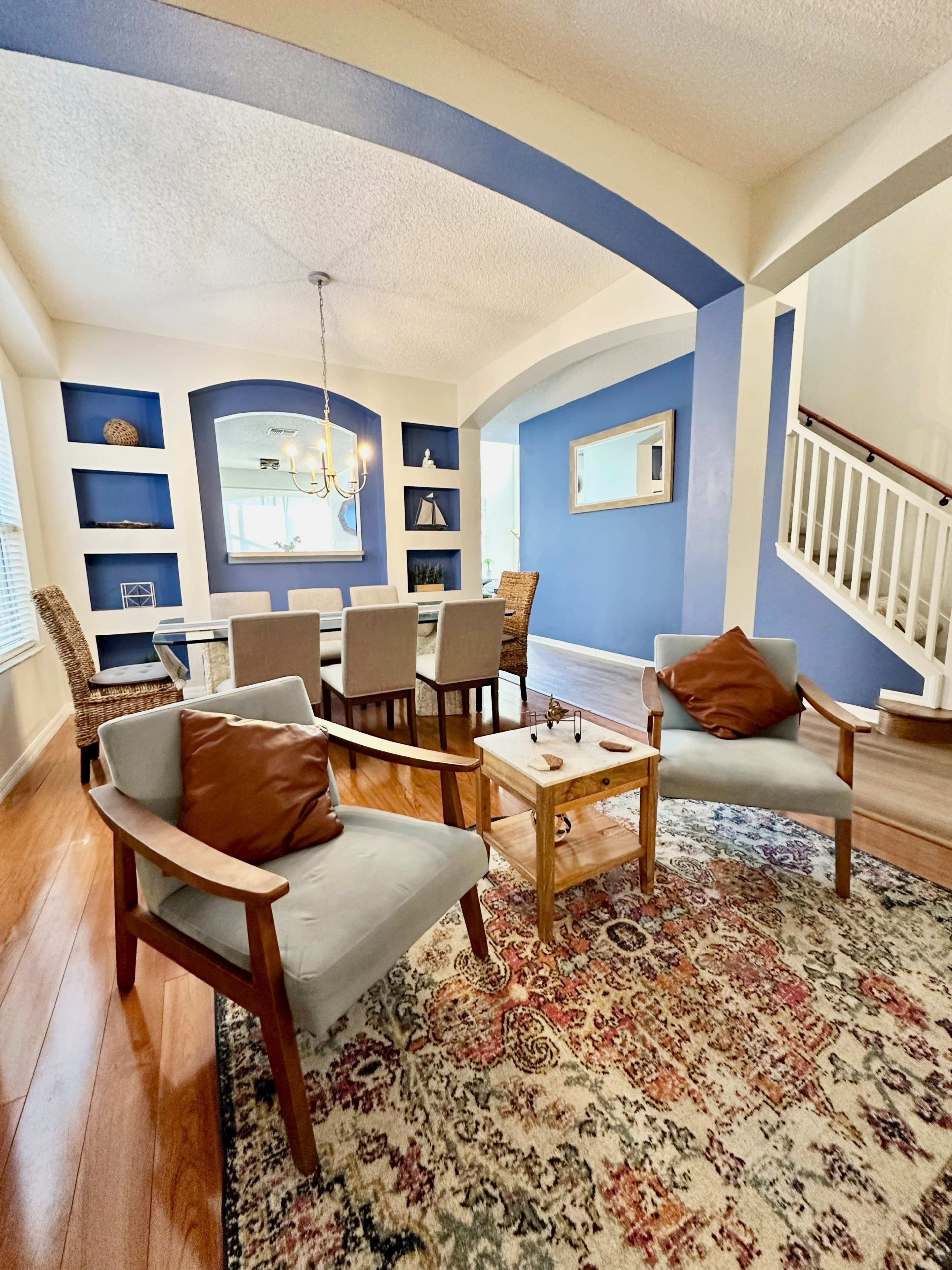 The image shows a cozy living and dining area featuring a blue accent wall, a wooden dining table, and seating with a mix of chairs and a patterned rug.