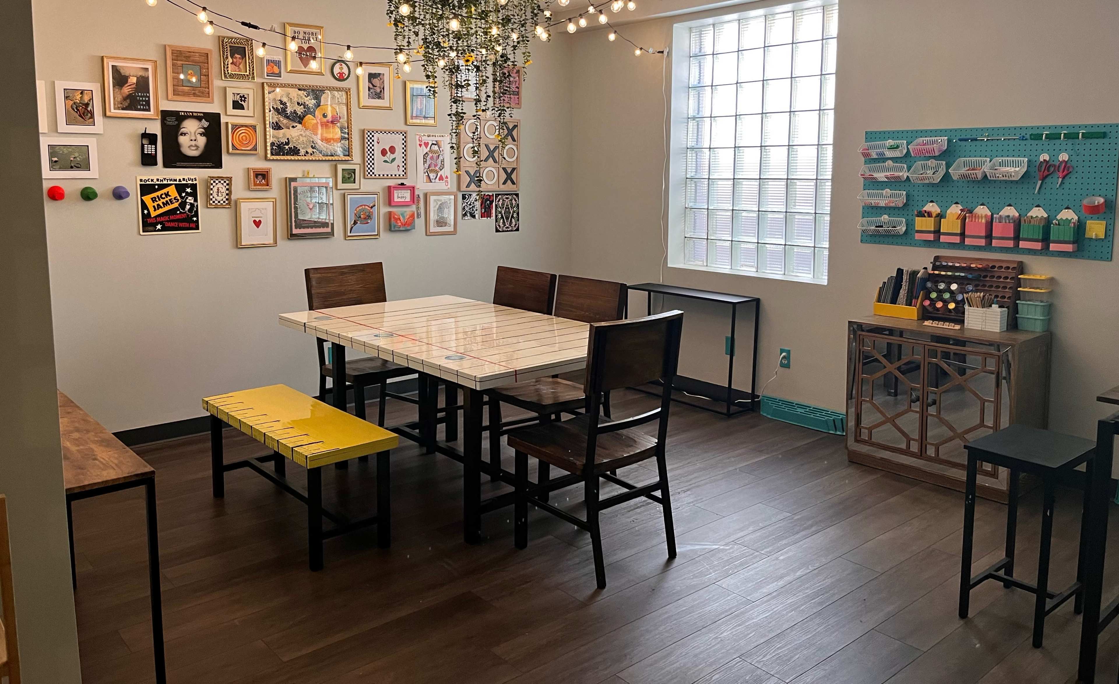 The room features a large table surrounded by chairs, with a wall adorned with various framed artworks and decorations, and strings of lights overhead.