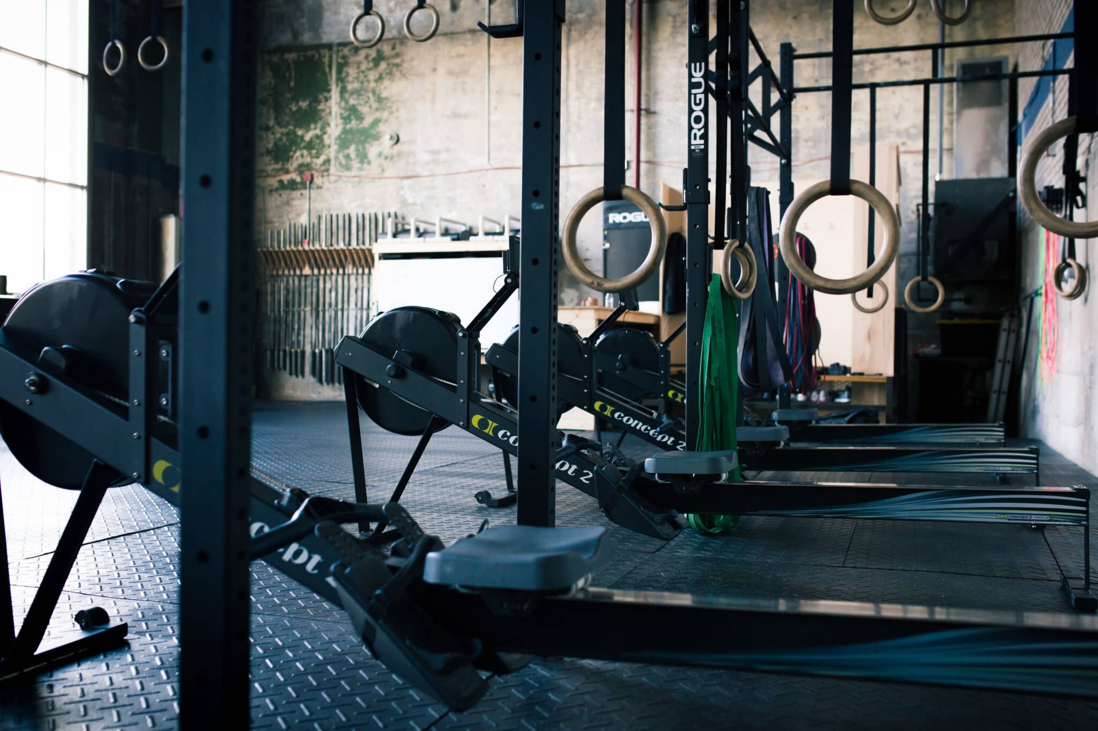The image shows a row of rowing machines set up in a gym with wooden rings hanging in the background.
