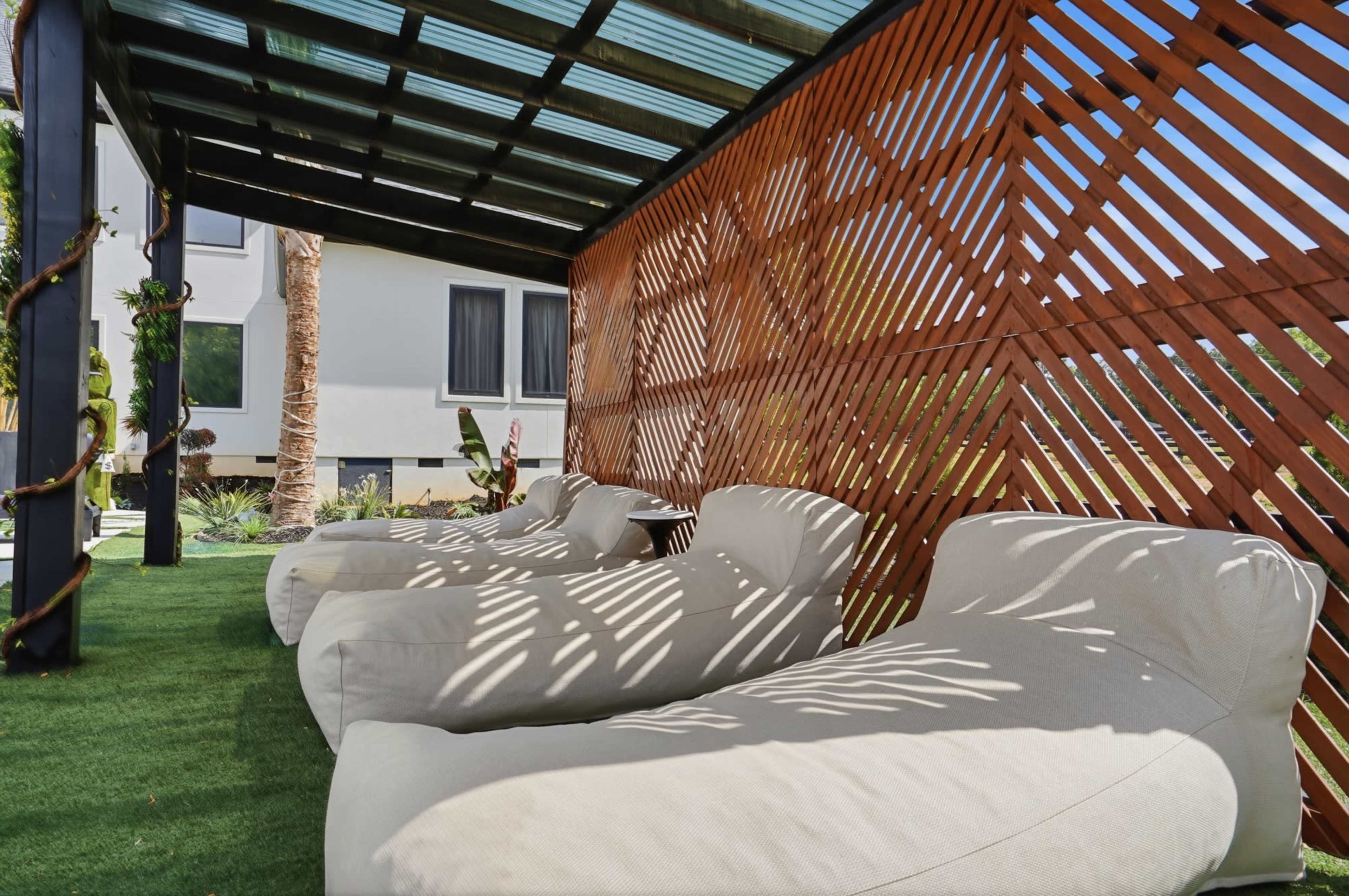 The image shows a shaded outdoor area with several white lounge chairs arranged on a grassy surface, with wooden slats creating a partial enclosure.