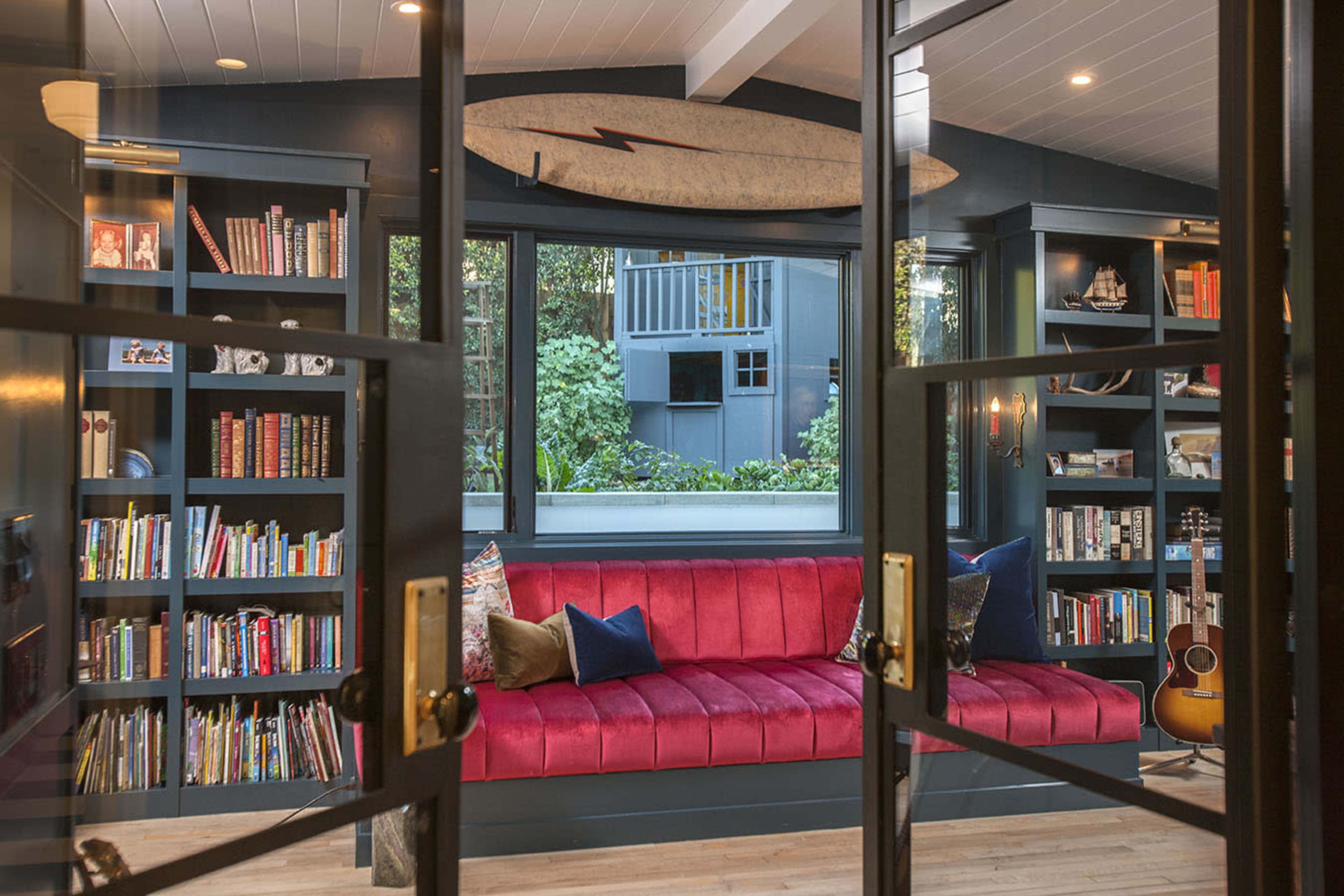 A cozy reading nook features a red sofa, built-in bookshelves filled with books, and a surfboard mounted above, set against large windows that overlook greenery.