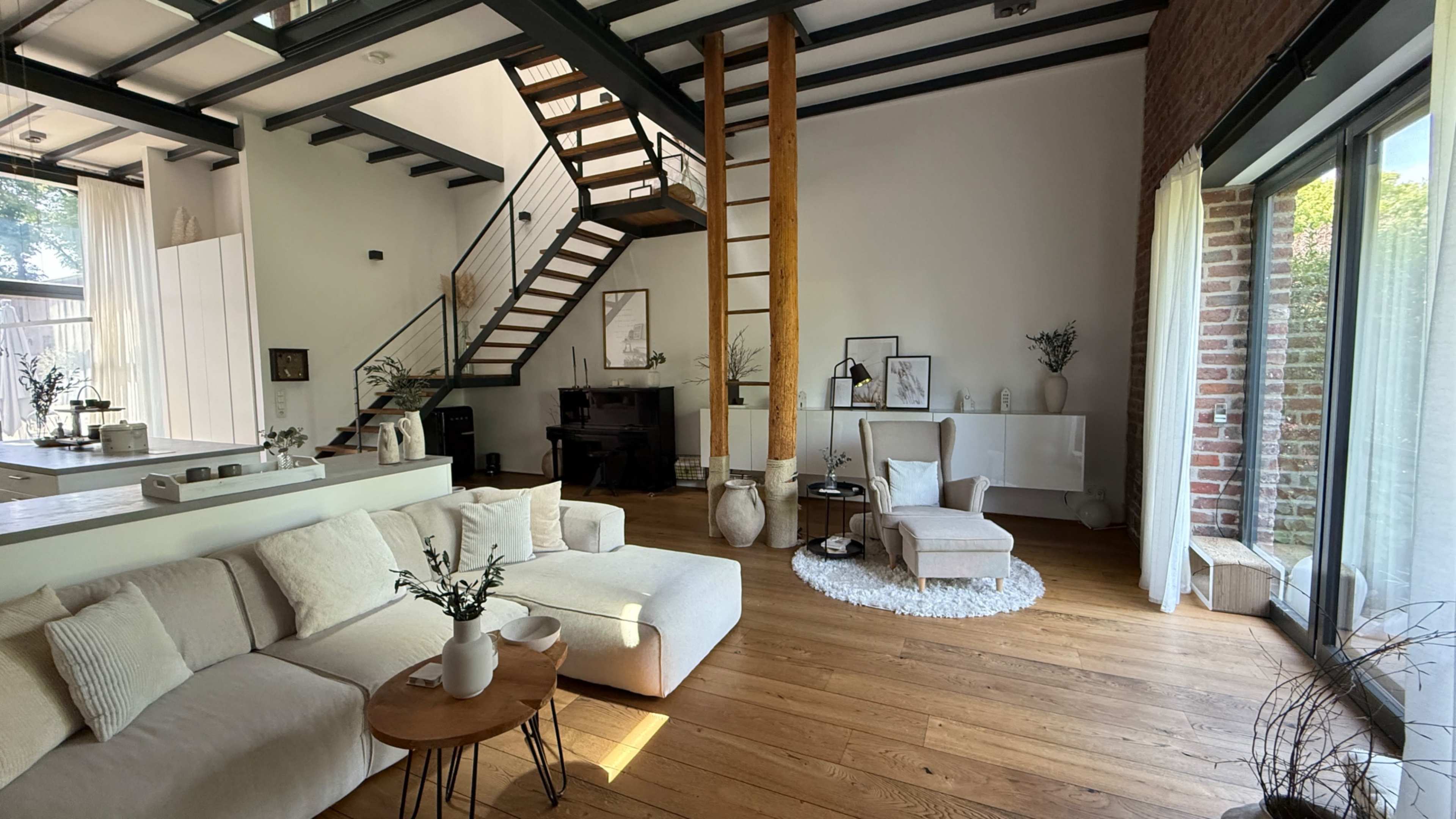 The image shows a modern living space with a staircase, a piano, and large windows that allow natural light to enter.