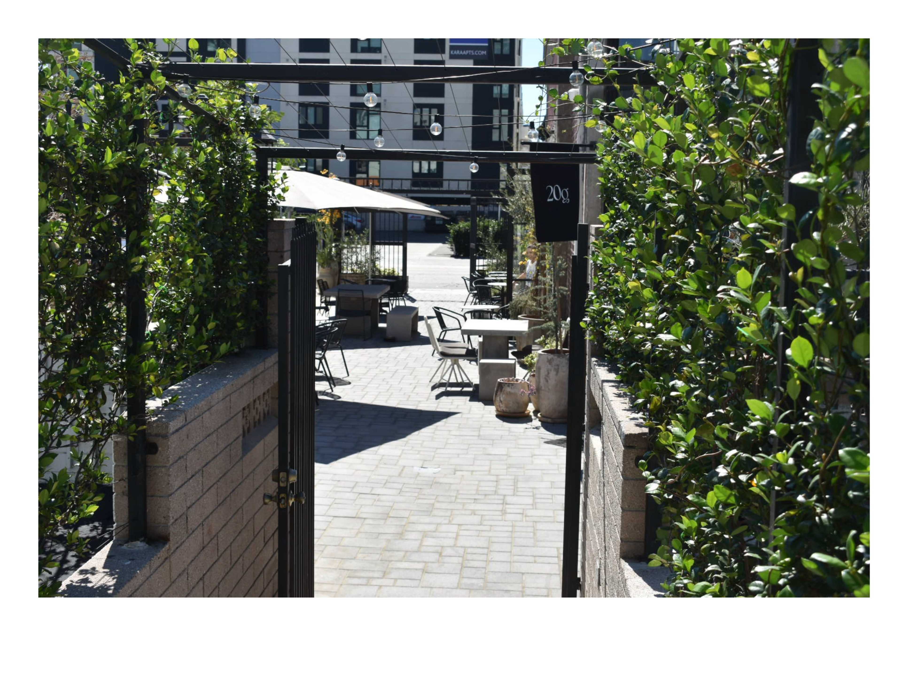 A paved pathway leads through a gate toward a seating area surrounded by greenery and outdoor tables.