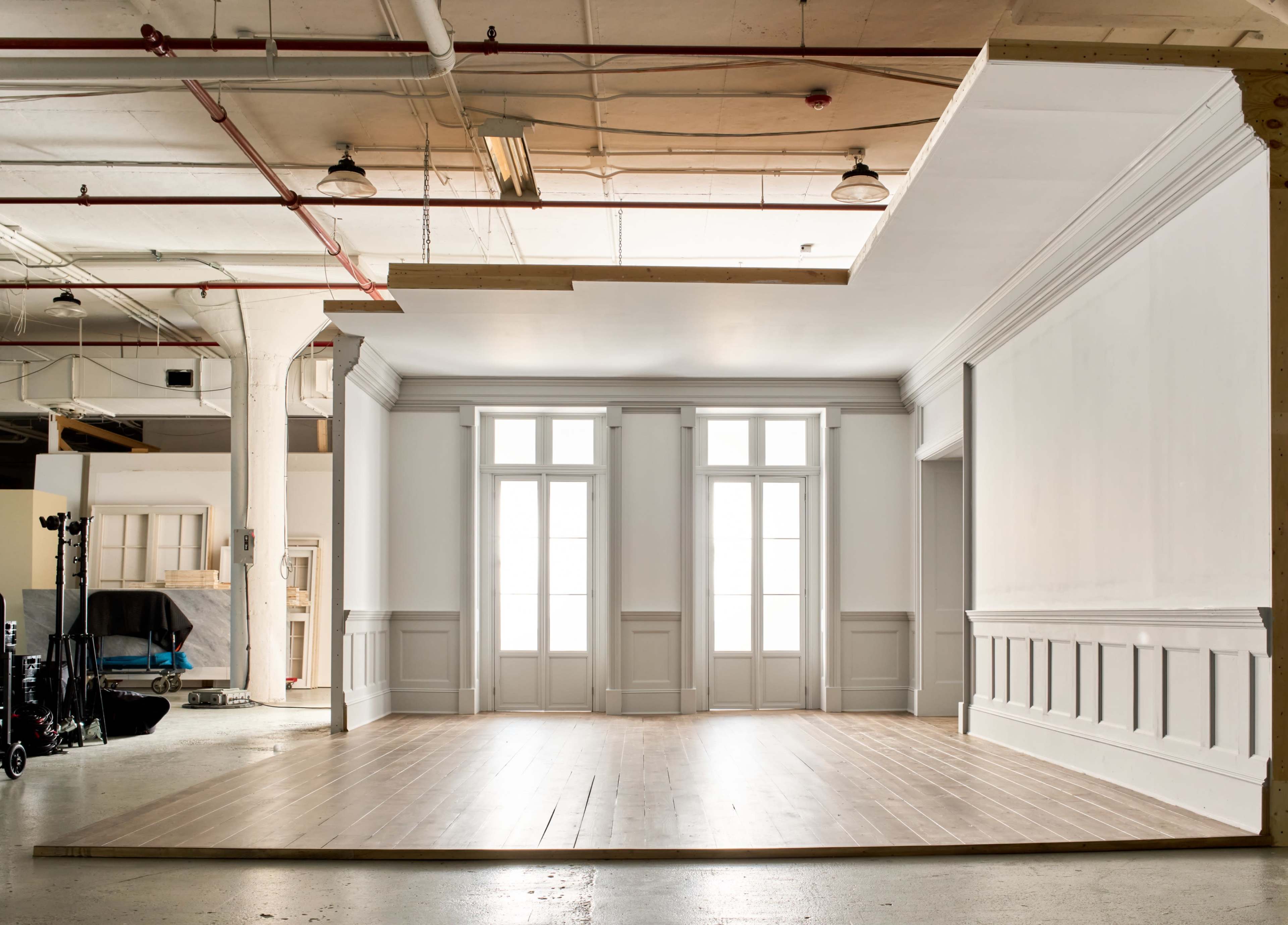 The image shows an empty studio space with wooden flooring and large windows, featuring white walls and a classic architectural design.