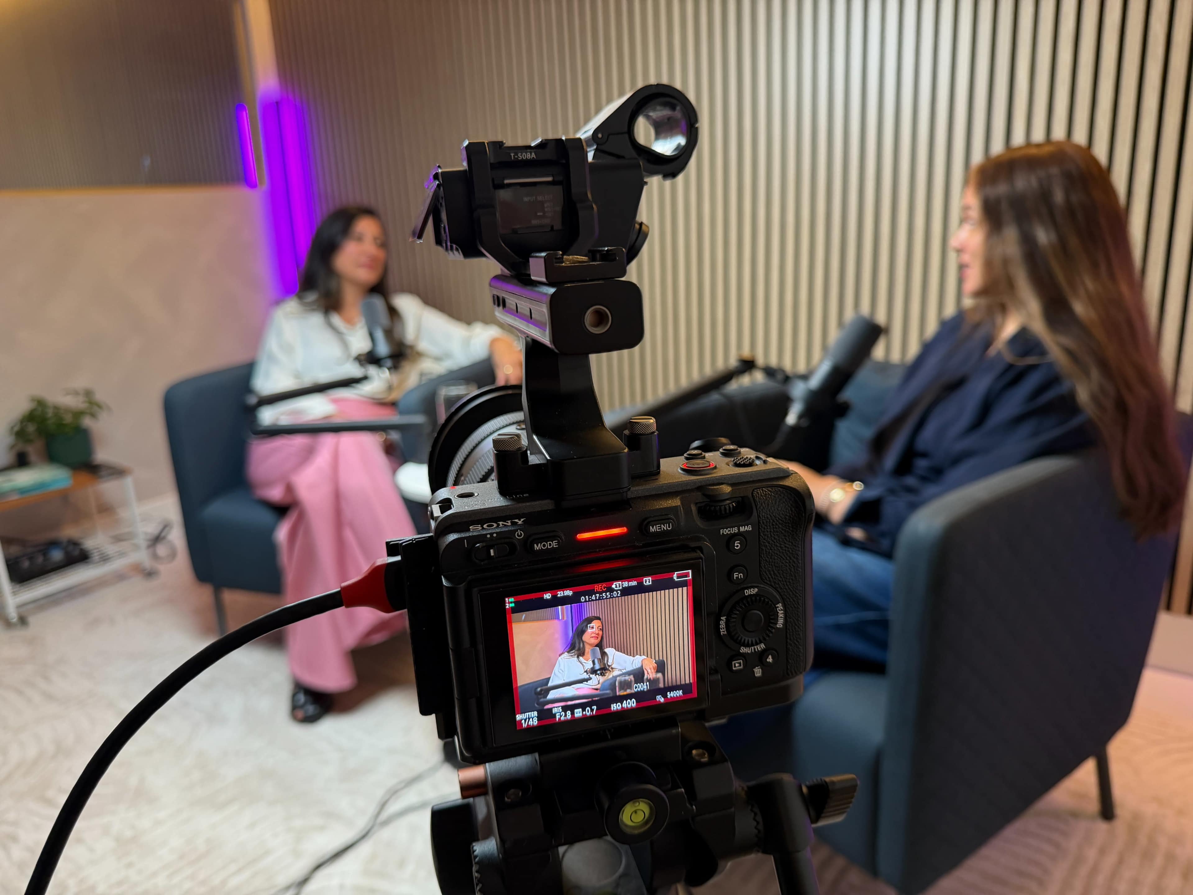 The image shows a camera set up in front of two women engaged in conversation during a podcast recording, with a blurred view of one woman in the foreground and the other visible on a screen in the background.