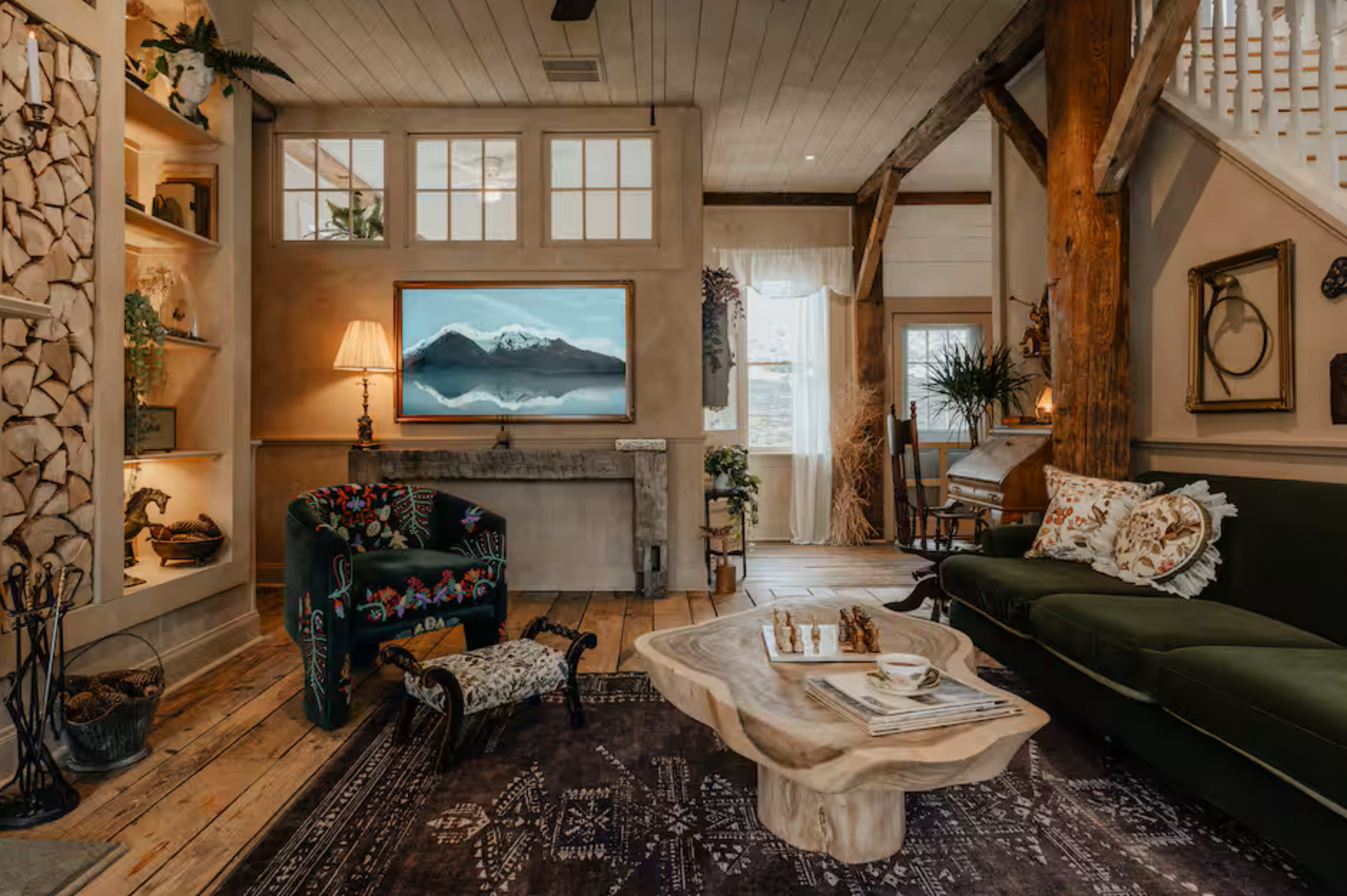The image shows a cozy living room featuring a green sofa, a decorative wooden coffee table, and a large television mounted on a wall with a mountain landscape displayed.