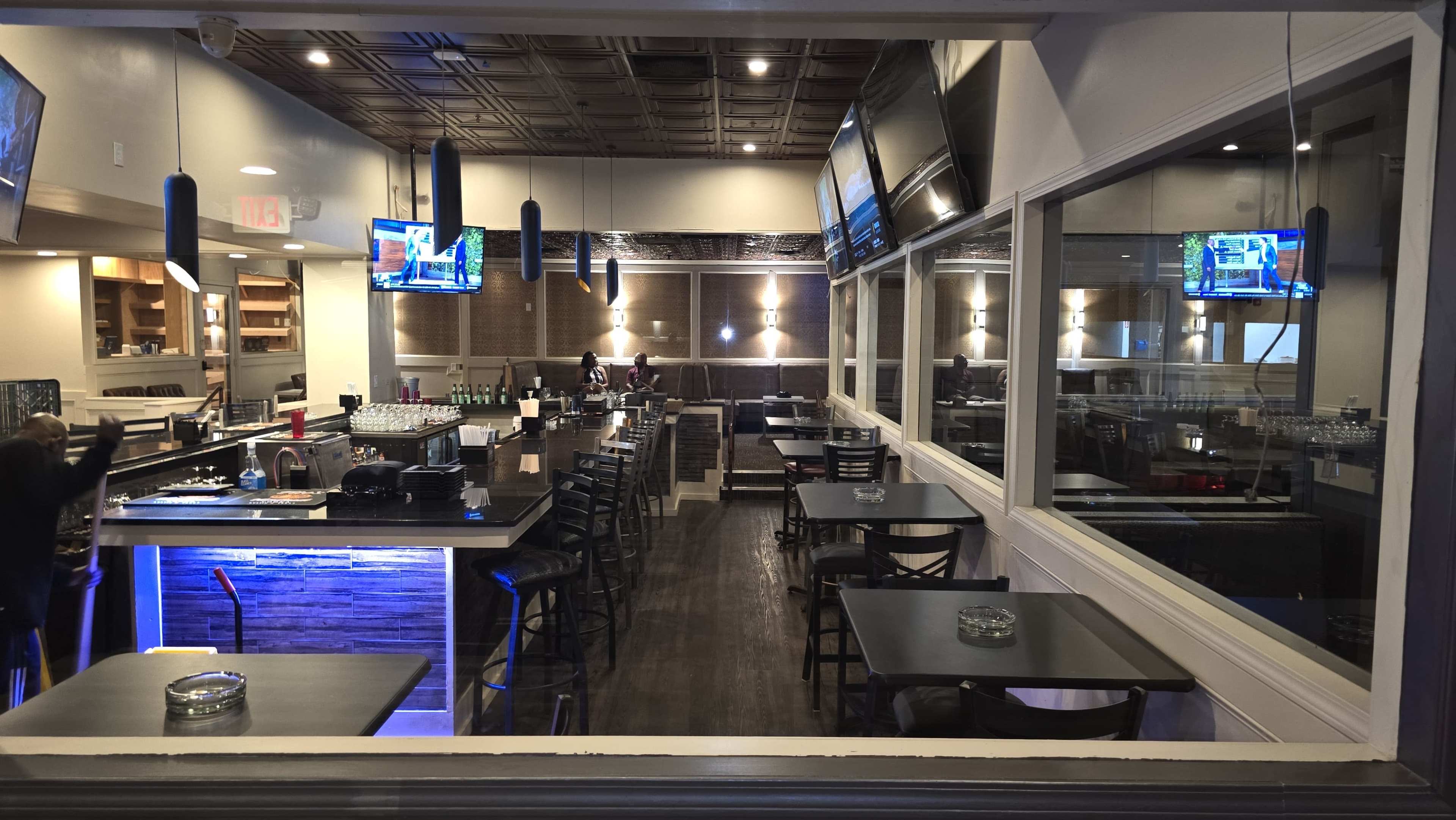 The image shows the interior of a modern restaurant with black tables and chairs, a bar with illuminated surfaces, and multiple television screens mounted on the walls.