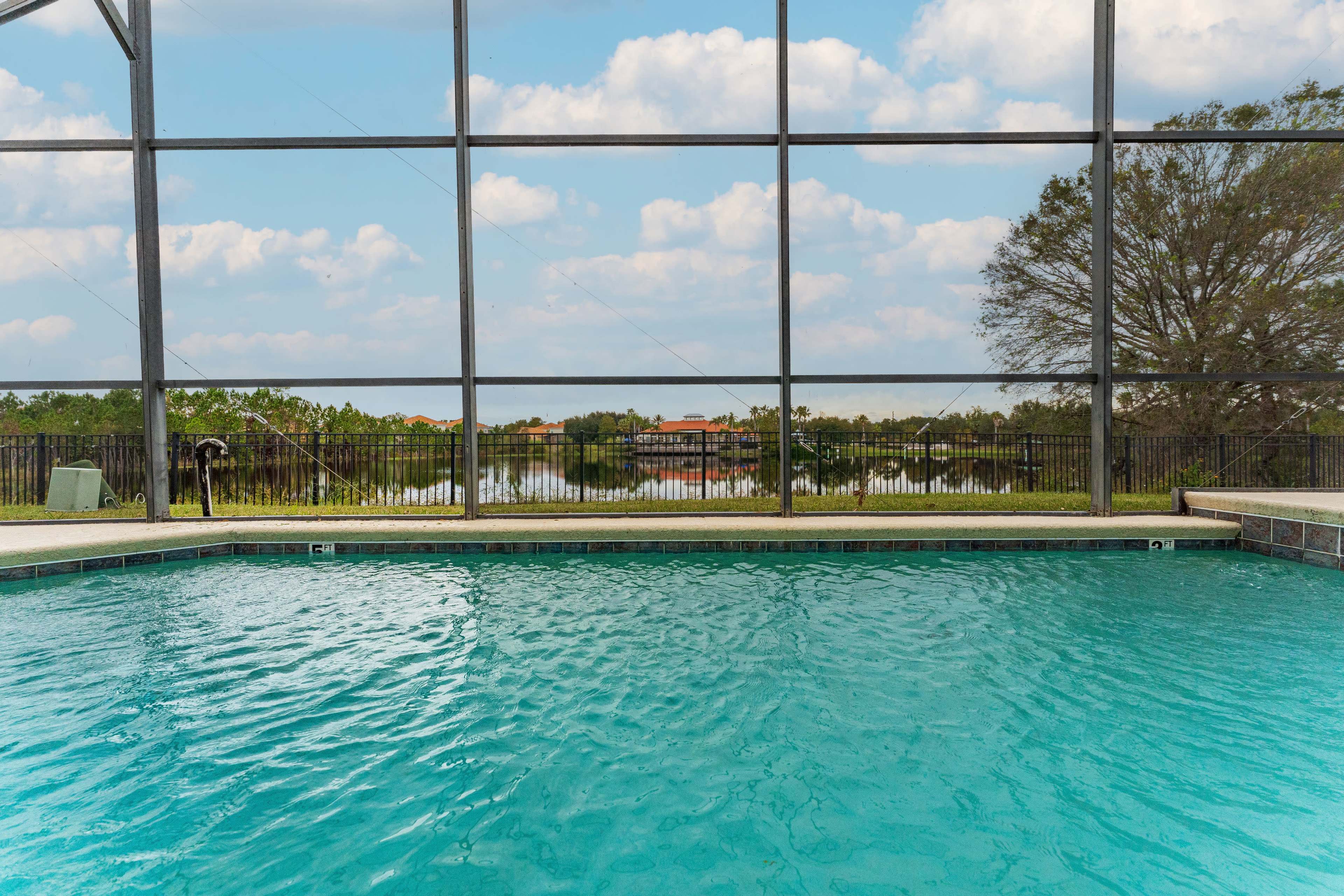A clear swimming pool is visible through a screened enclosure, with a tranquil view of trees and a lake in the background.