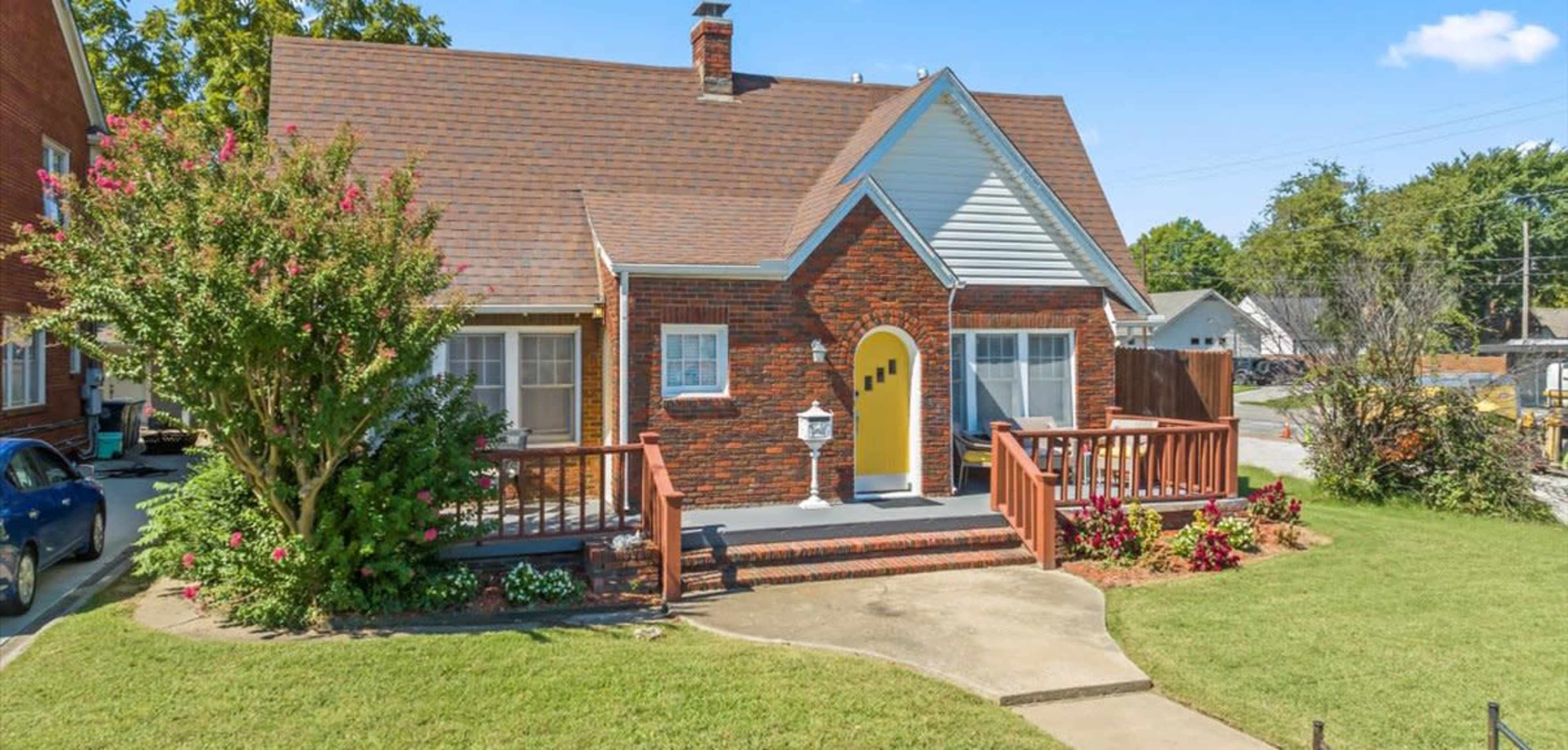 A brick house with a gabled roof features a yellow door and a front porch surrounded by landscaping.