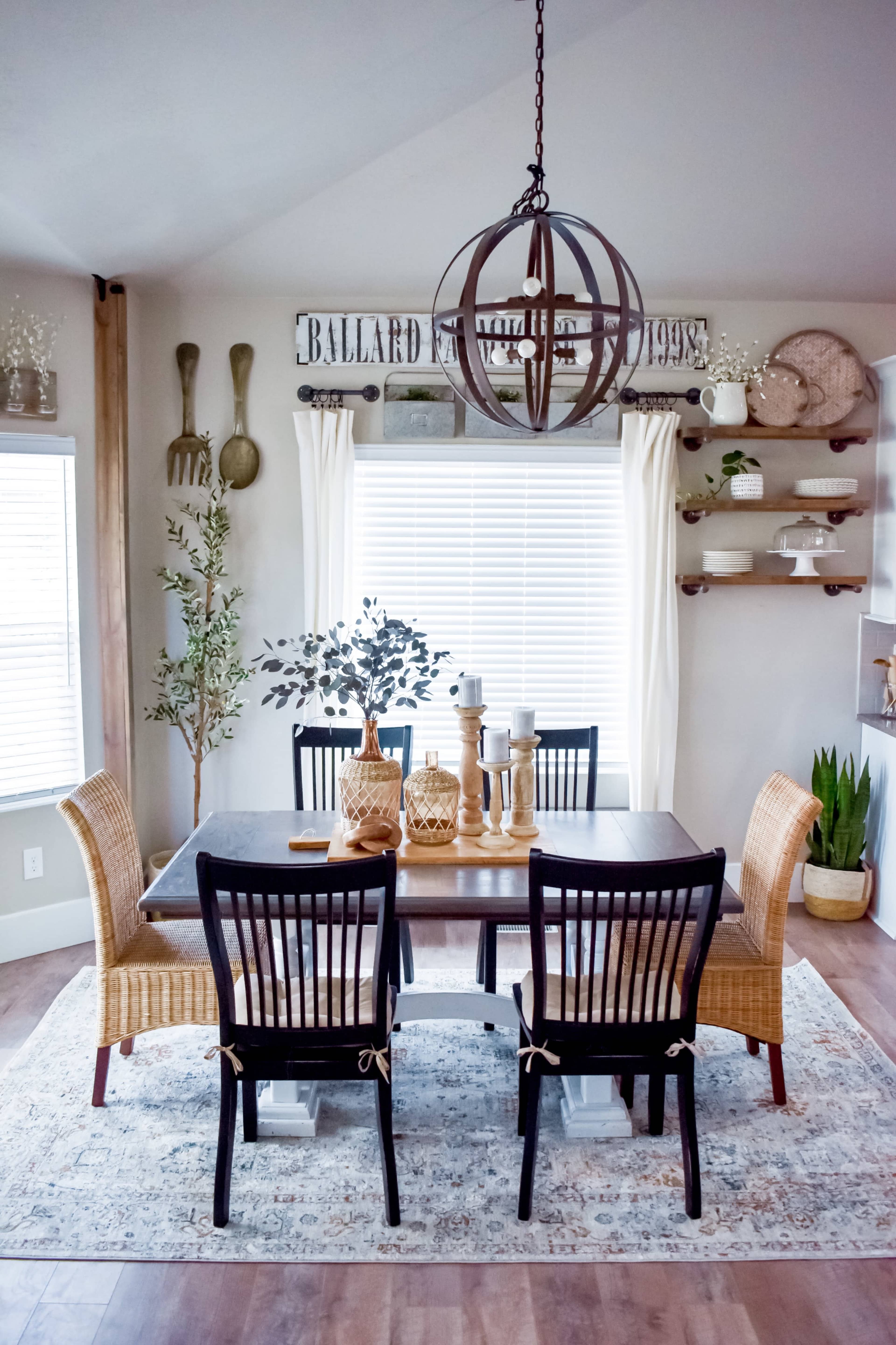 A dining room features a wooden table surrounded by black and wicker chairs, with a large spherical light fixture above and decorative items on shelves in the background.