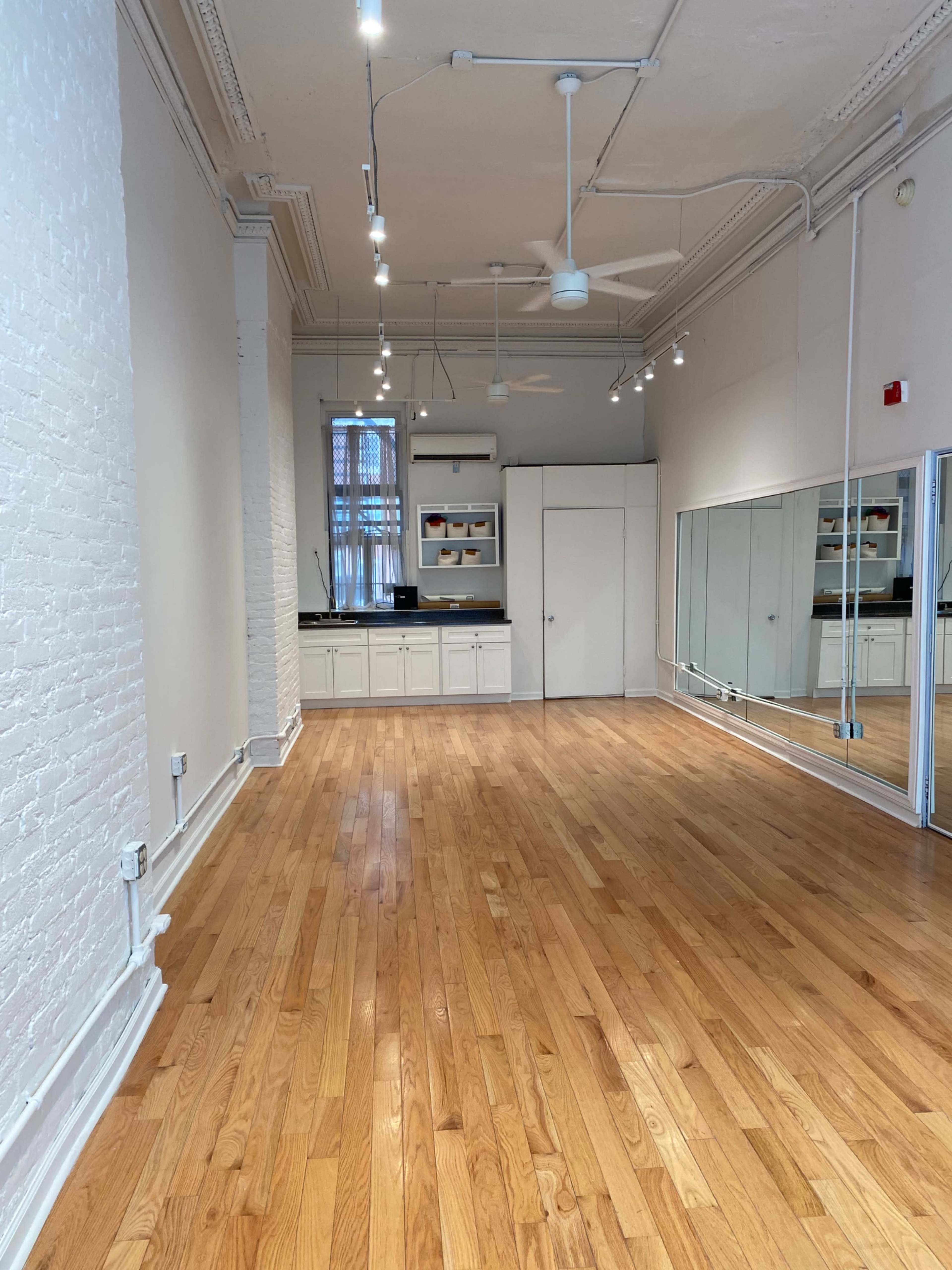 A well-lit, empty room with hardwood flooring, a large mirror on one wall, and a kitchenette area.