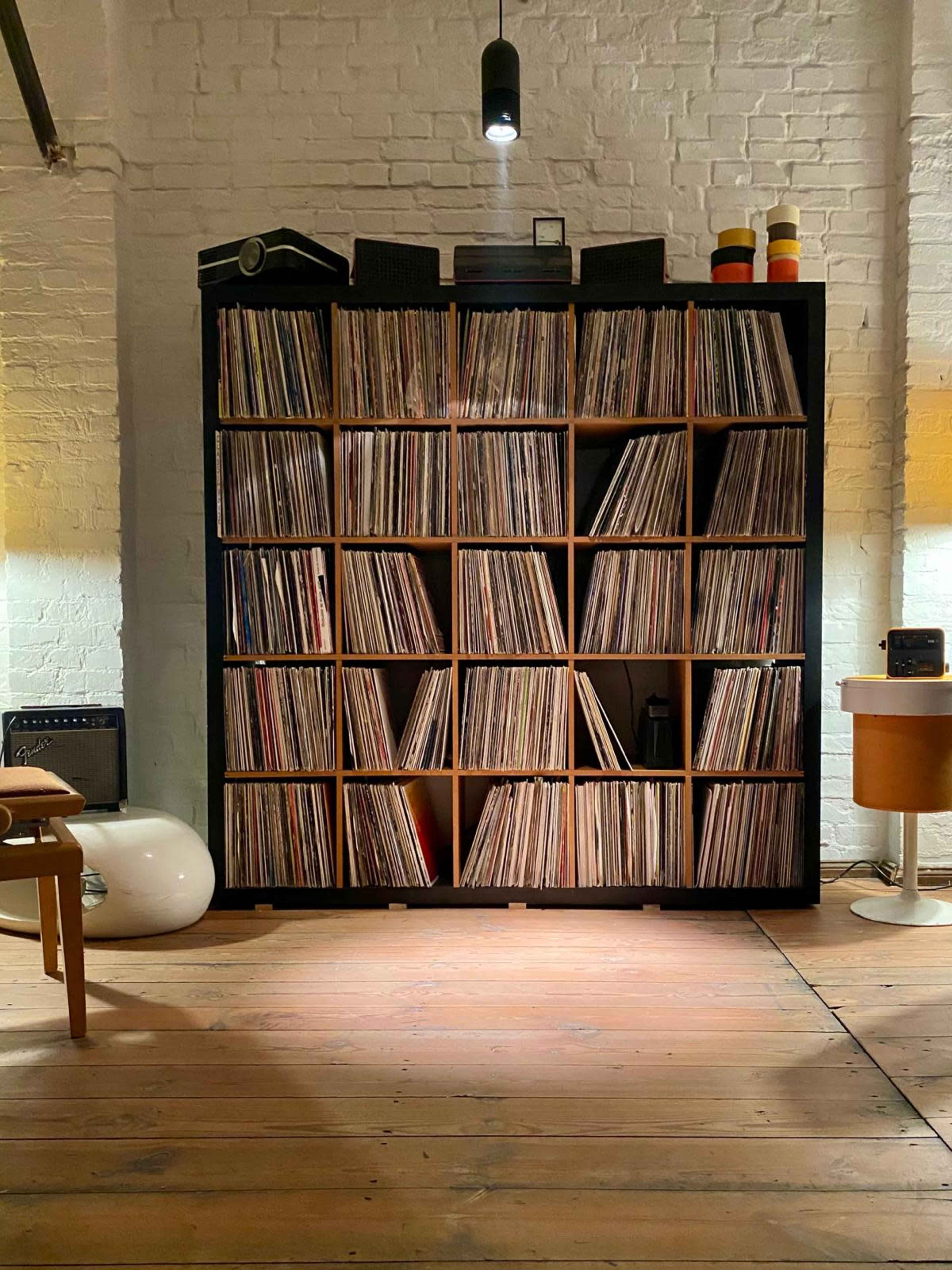 A wooden shelving unit filled with vinyl records is positioned against a white brick wall in a room with wooden flooring.