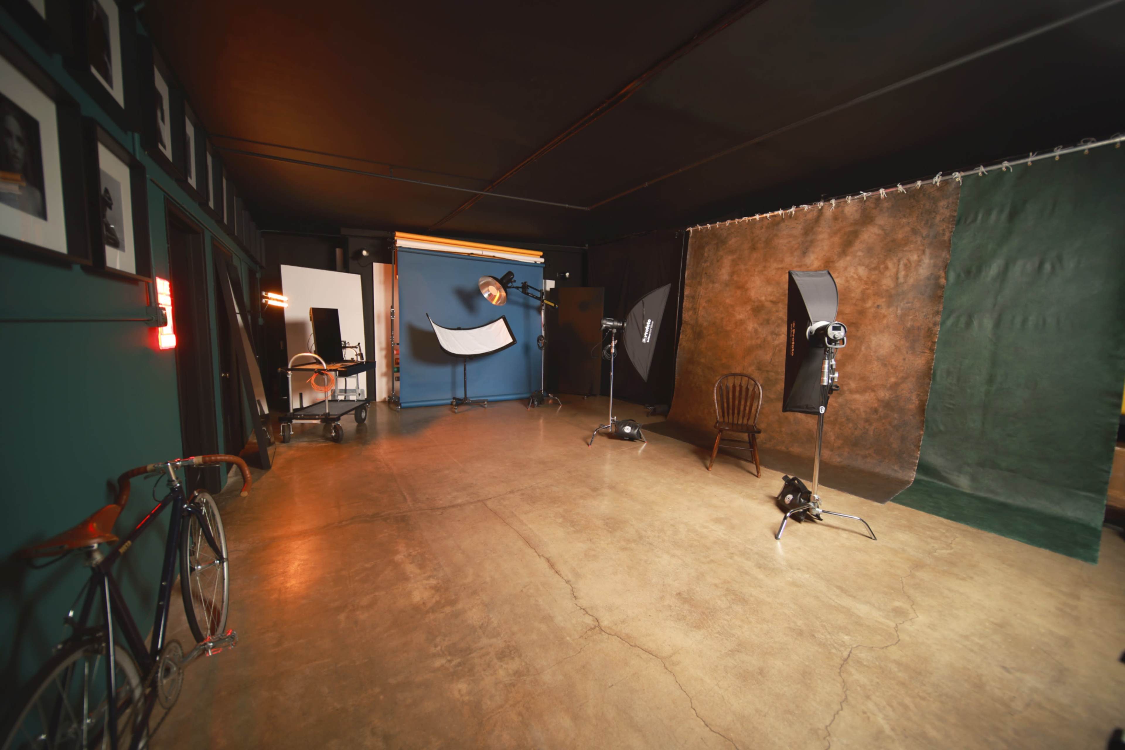 The image depicts a photography studio with colorful backdrops, lighting equipment, and a wooden chair, all arranged on a polished concrete floor.